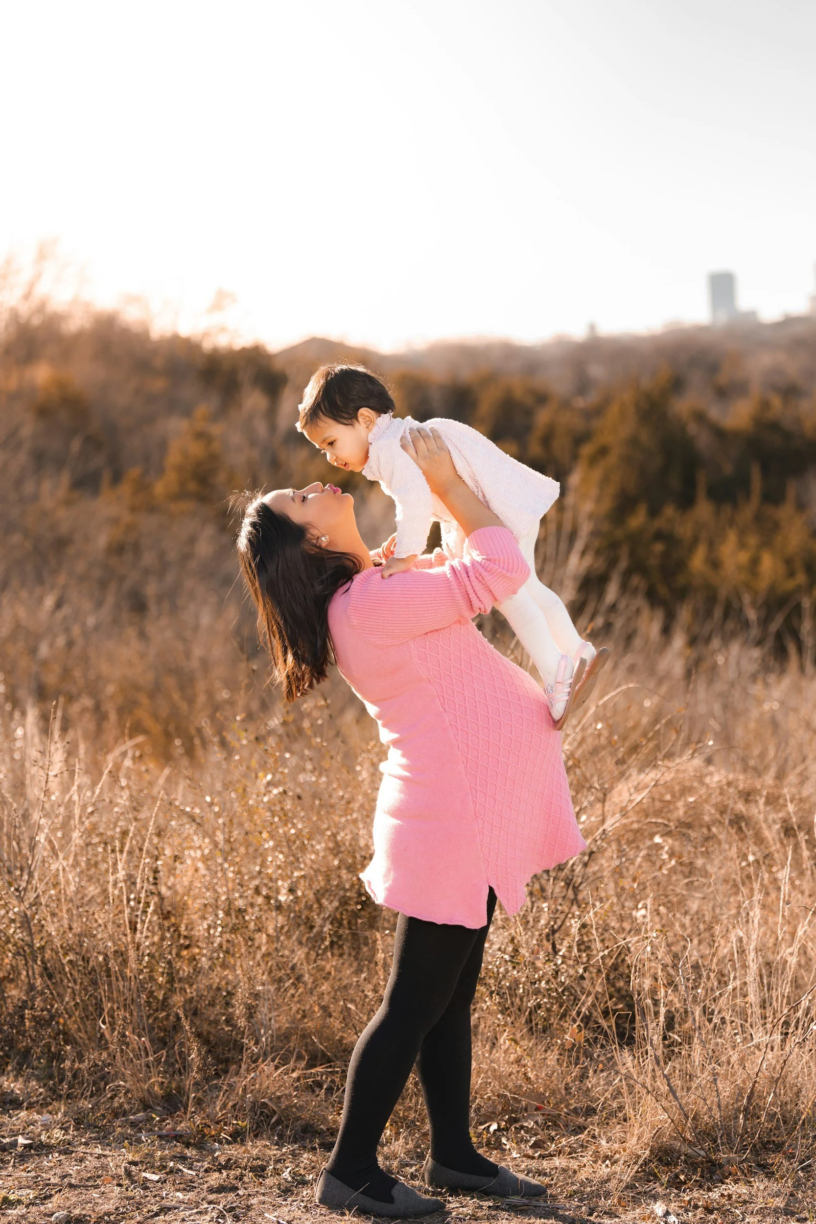 Pregnant mother holding her toddler in the air and giving a kiss.
