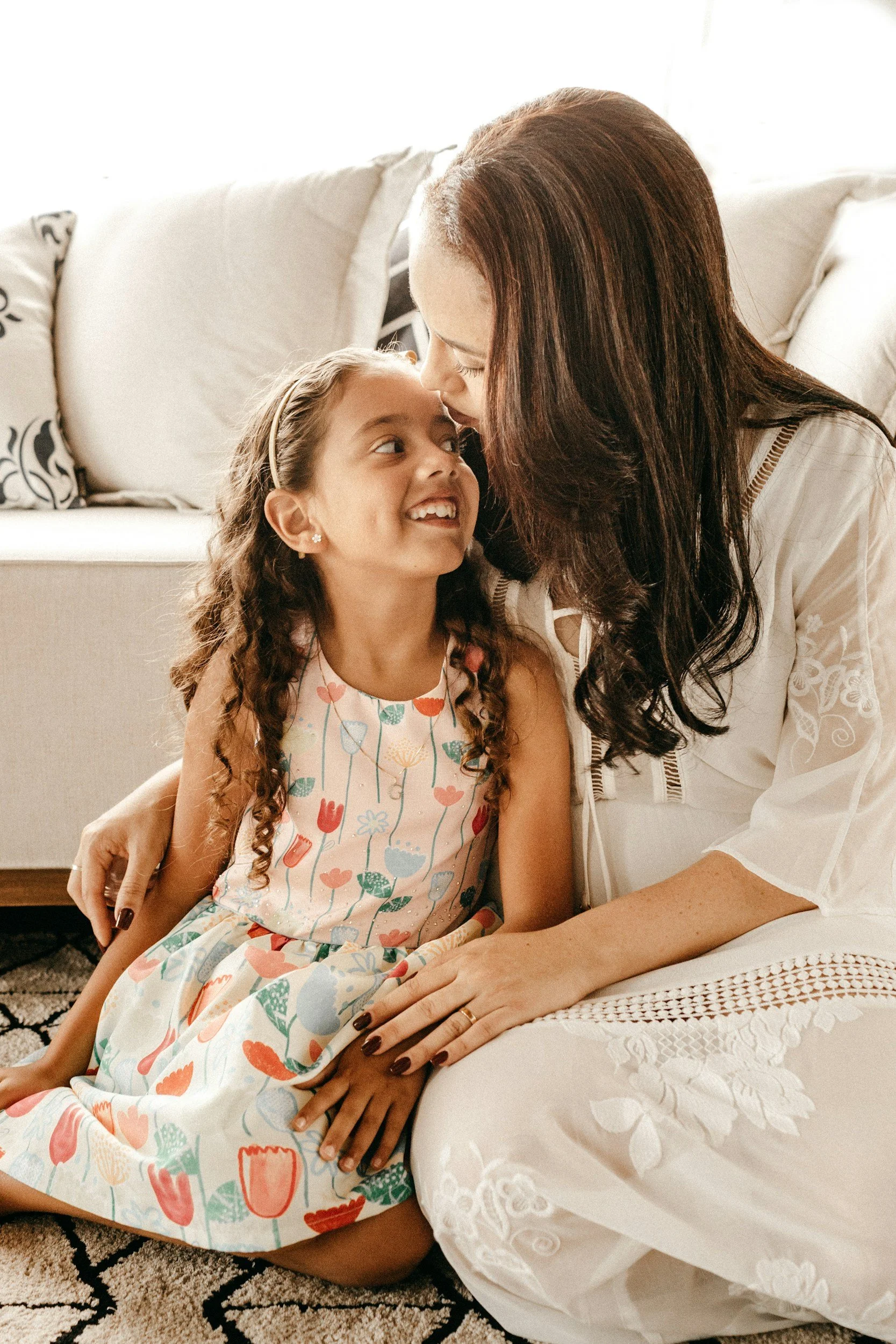 A mother sitting on the floor with her young child, tenderly kissing the child's forehead to offer comfort and somatic regulation.