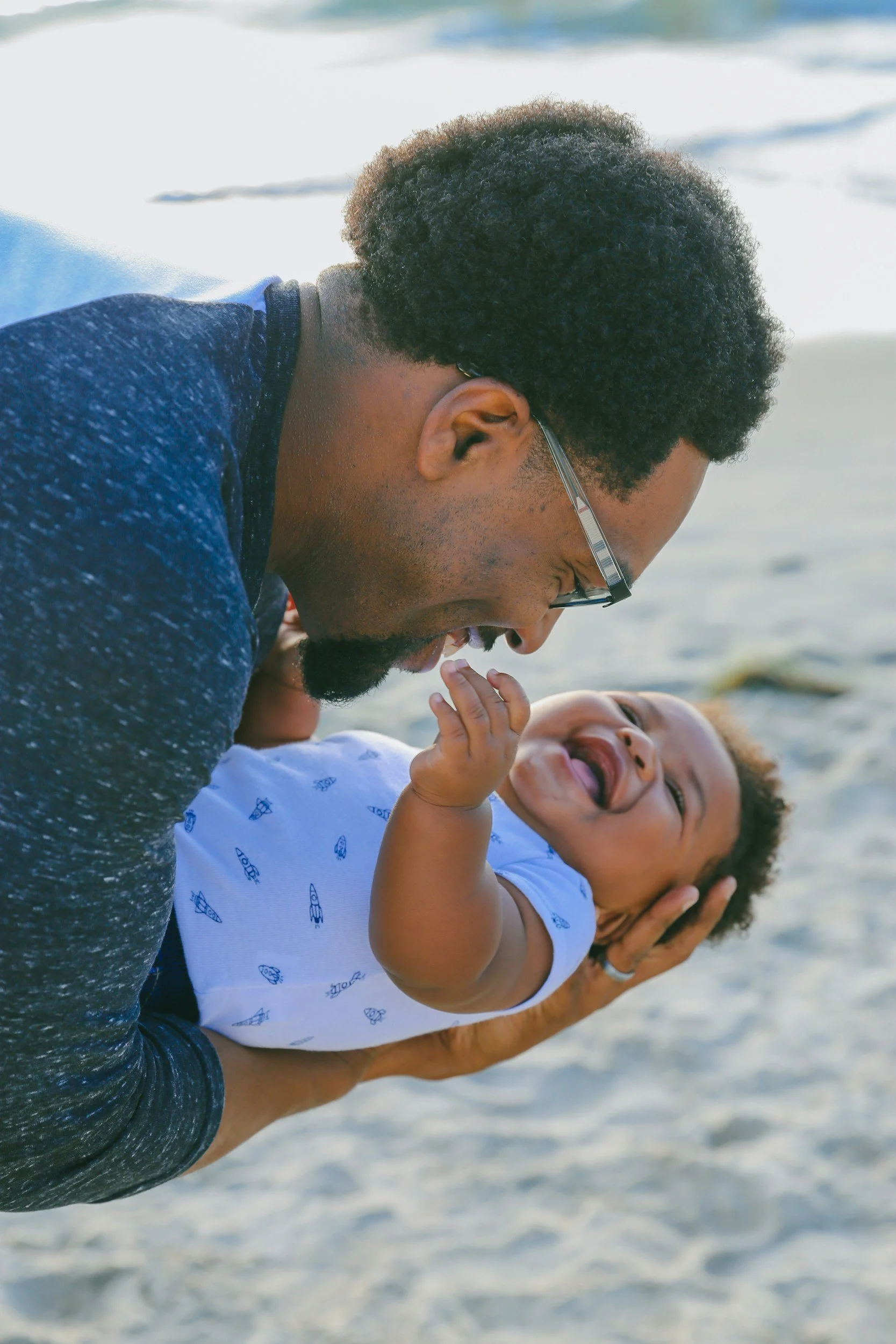 A laughing father holding his baby up close to his face while engaging in joyful, interactive play inside a South Bay family home.