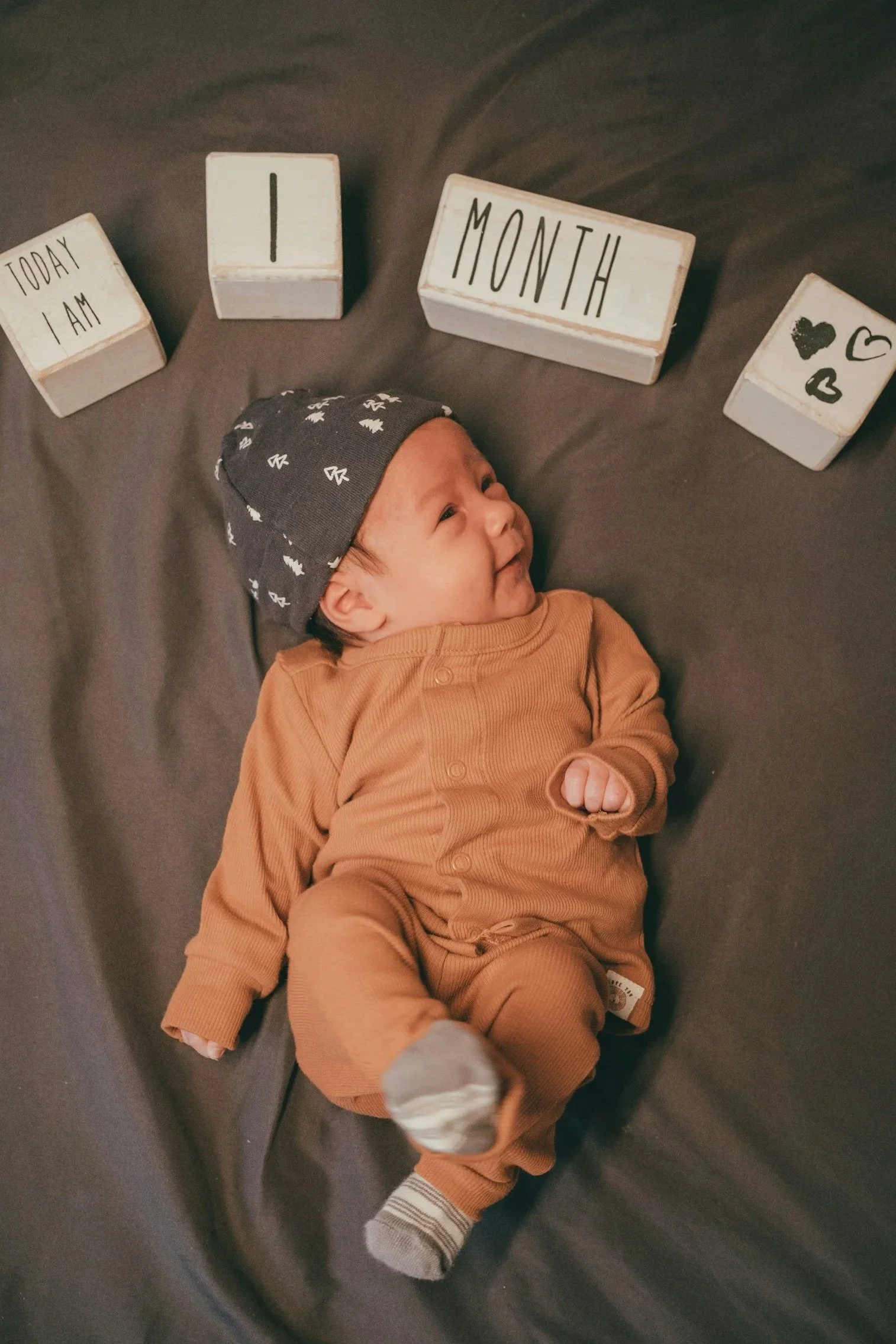 Baby lying on a dark gray surface, wearing a brown outfit, gray hat with small white designs, and gray socks. Baby is progressing in developmental milestones.