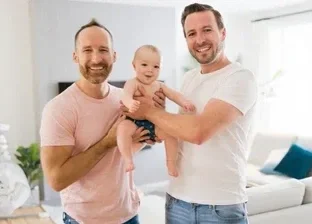 Two fathers posing together while holding their baby facing forward toward the camera, highlighting diverse family structures in early childhood wellness.