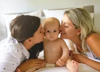 Two mothers simultaneously kissing their baby’s cheeks, showcasing responsive co-regulation and emotional attunement in a queer-affirming home.
