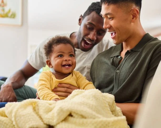 A Black father and an Asian father smiling warmly while holding their infant together, demonstrating inclusive early childhood attachment bonds.