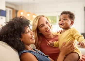 Two mothers sitting closely on a living room couch, smiling and holding their infant baby together in a safe, nurturing home environment.