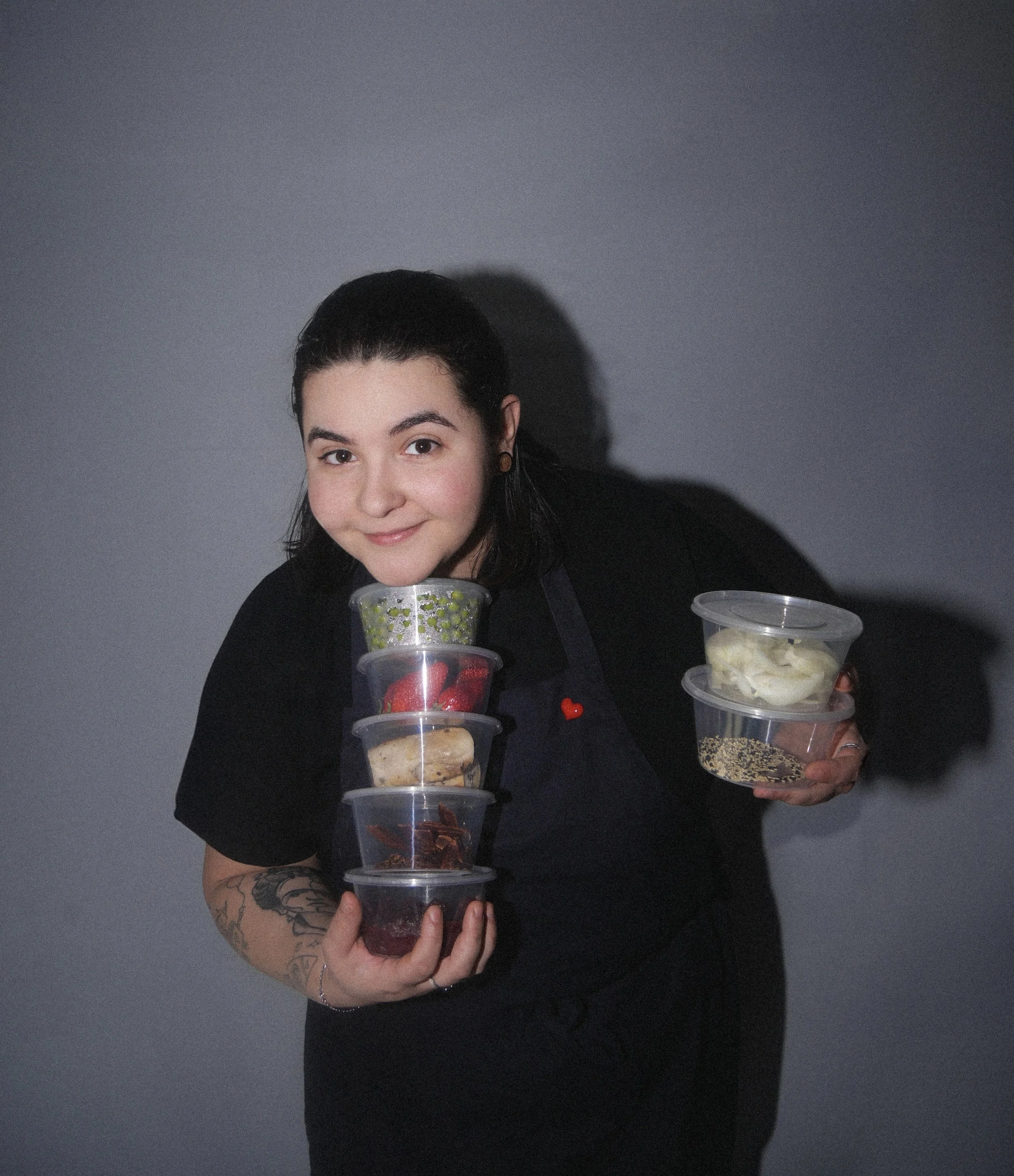 Femme tenant plusieurs contenants en plastique avec des aliments, souriant, portant un tablier noir, contre un mur gris.