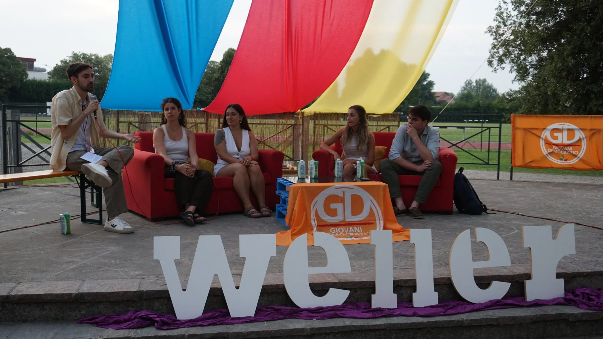 Un gruppo di giovani partecipanti a un dibattito seduti su un divano rosso sotto una grande tenda colorata, con un moderatore che tiene un microfono e una scritta 'we' in primo piano. Sullo sfondo si vede un campo sportivo e un banner con il logo del partito Giovanile Democratico.