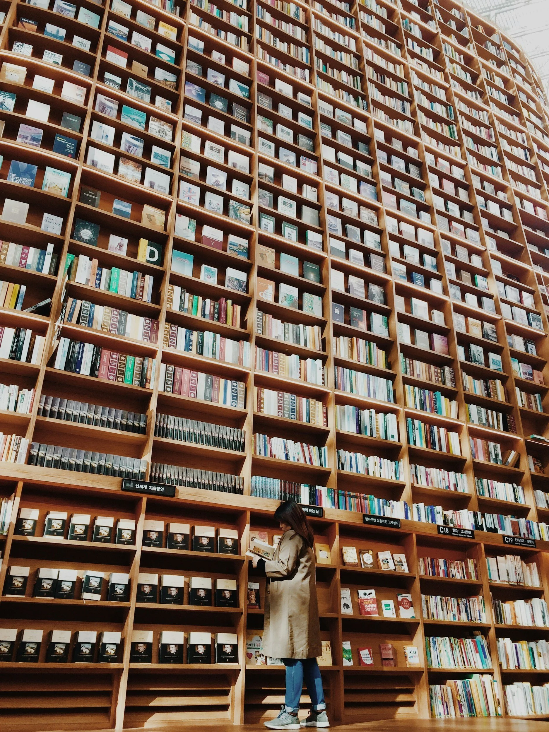 A woman wearing a beige coat, blue jeans, and sneakers standing at a bookshelf in a large library, browsing through books.