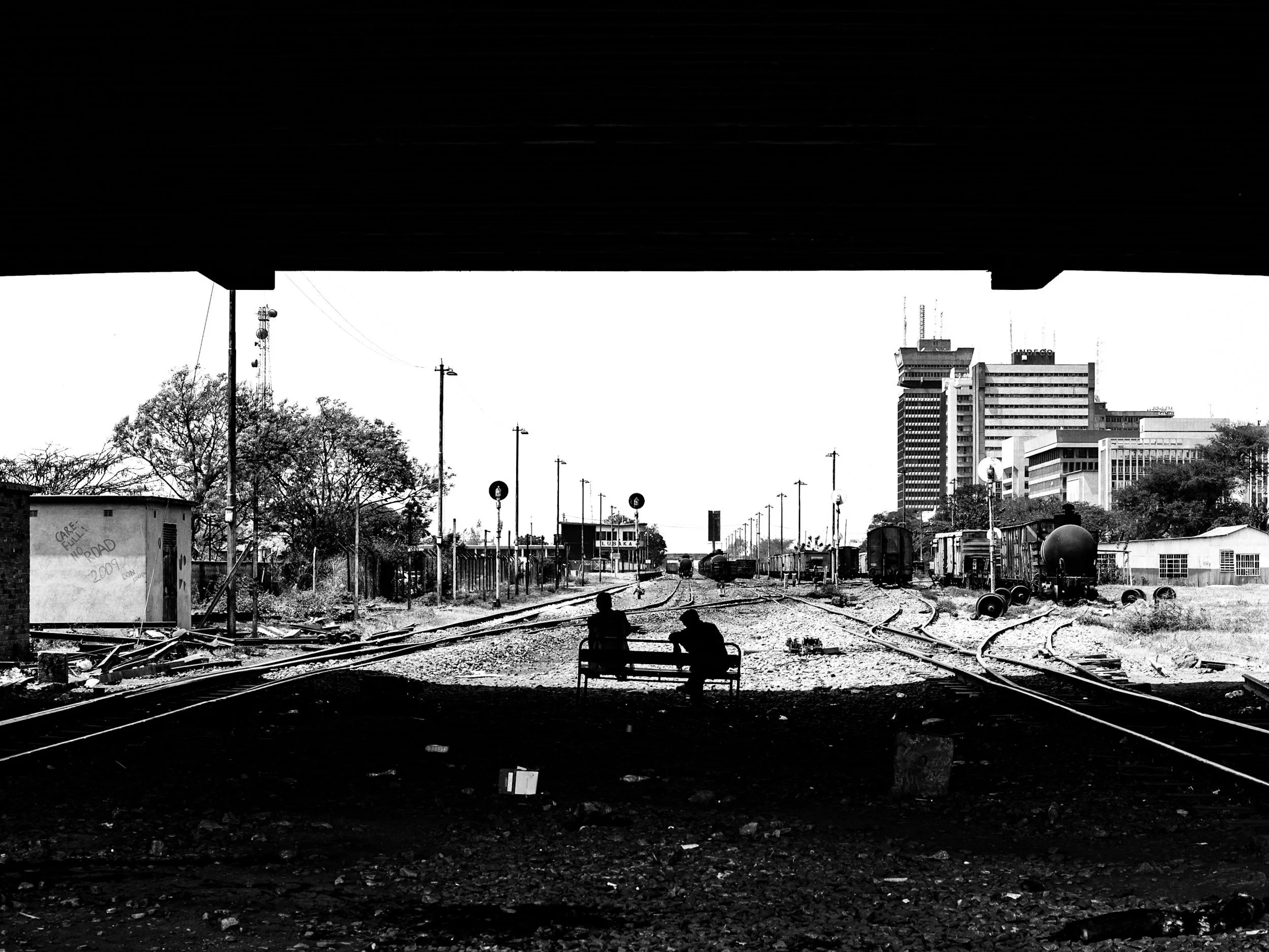 Black and white photo of two people sitting on a park bench under a bridge, with railway tracks and industrial buildings and high-rise buildings in the background.