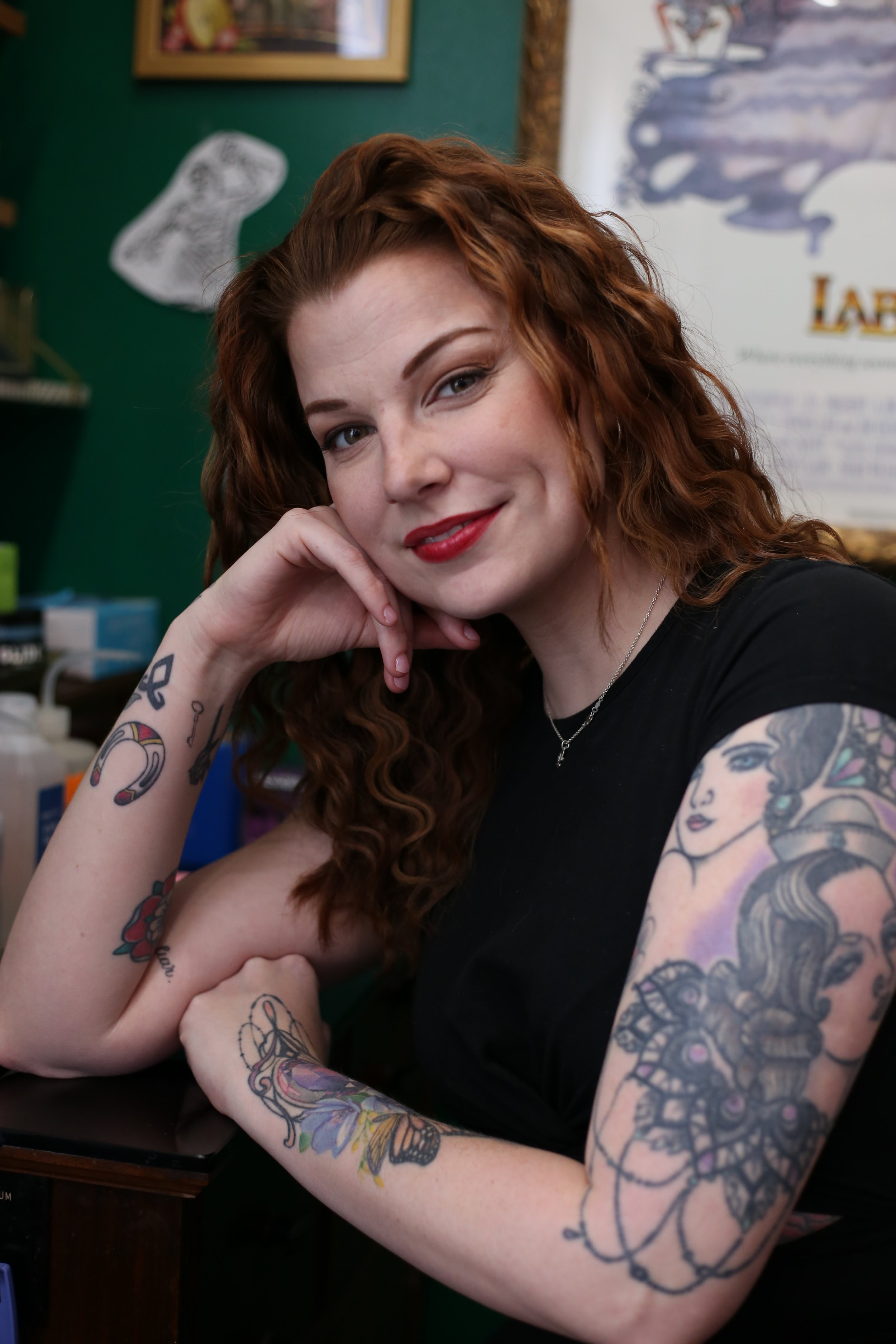 Young woman with red curly hair and tattoos on her arms, smiling and resting her chin on her hand, sitting at a table in a room with green walls, a wall art piece, and framed pictures.