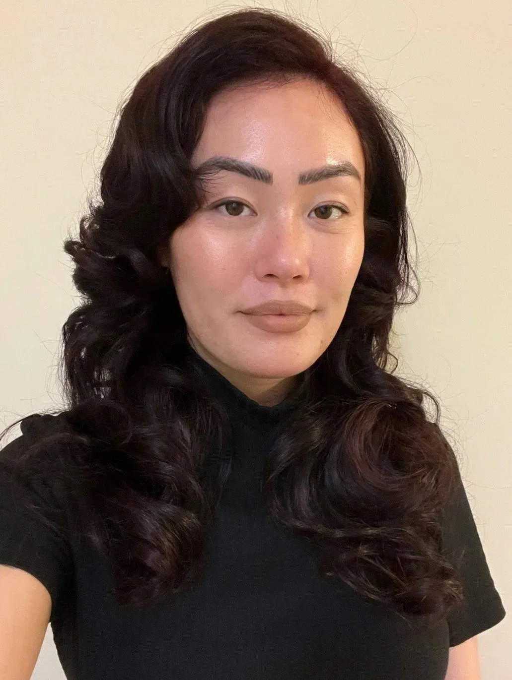 A woman with shoulder-length dark brown wavy hair, wearing a black turtleneck, looking at the camera with a neutral expression against a plain light-colored background.