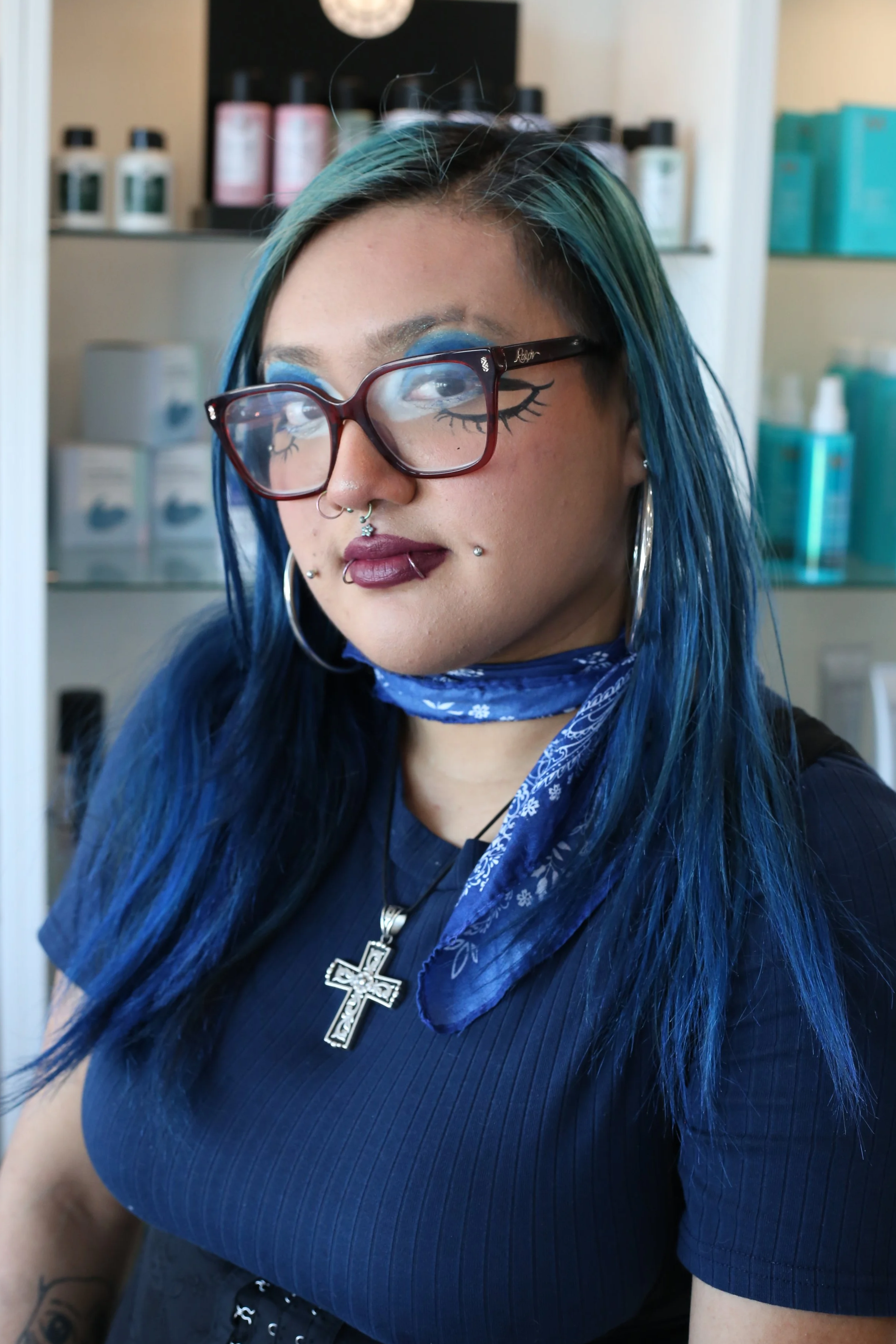 A woman with blue hair, glasses with eyelash illustrations, and multiple facial piercings wearing a blue bandana and jewelry, inside a store or salon.
