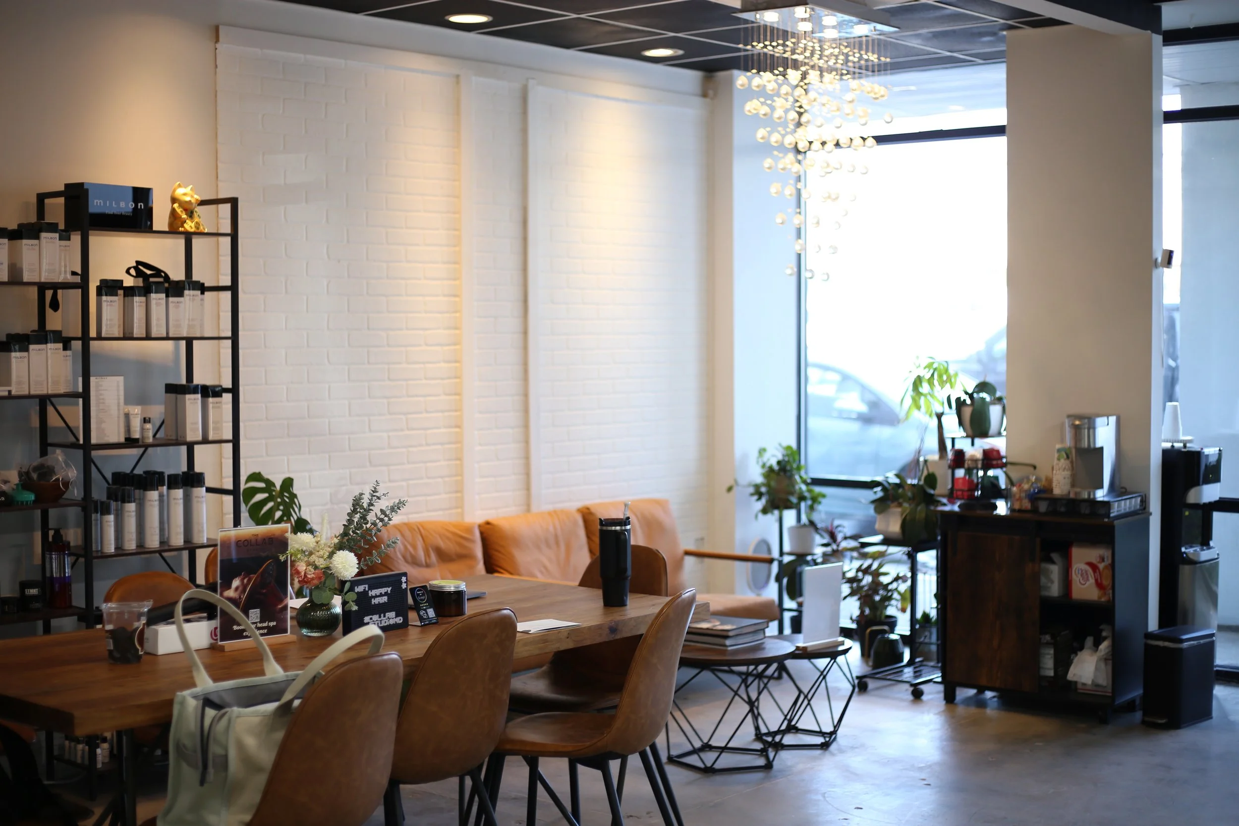 Interior of a cozy café with a wooden table, leather chairs, potted plants near large windows, white brick wall, and shelves with product bottles and decorative items.