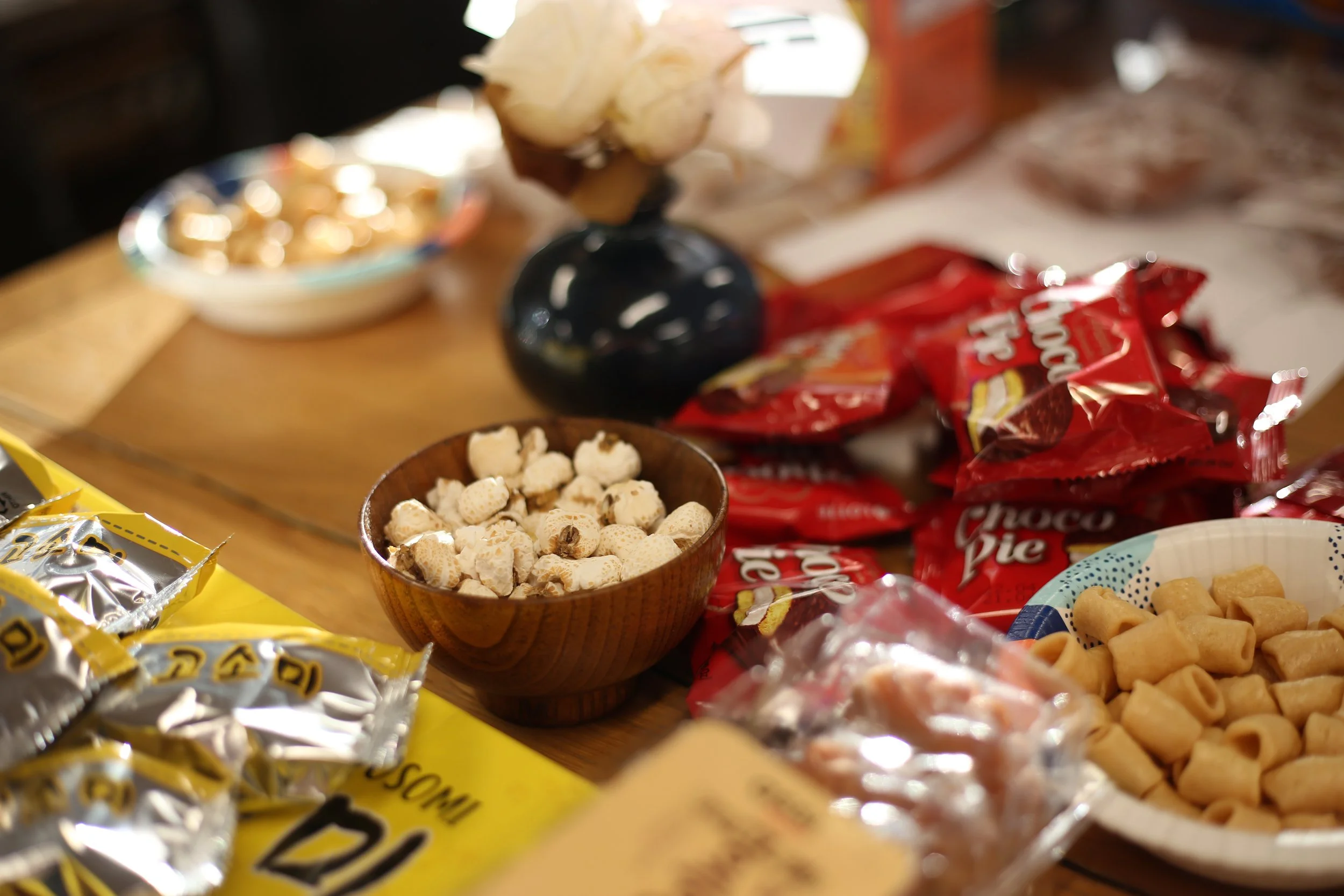 Assorted snacks and candies on a wooden table, including small white popcorn in a wooden bowl, red snack bags, a white bowl of bite-sized crackers, and a black vase with a cotton flower.