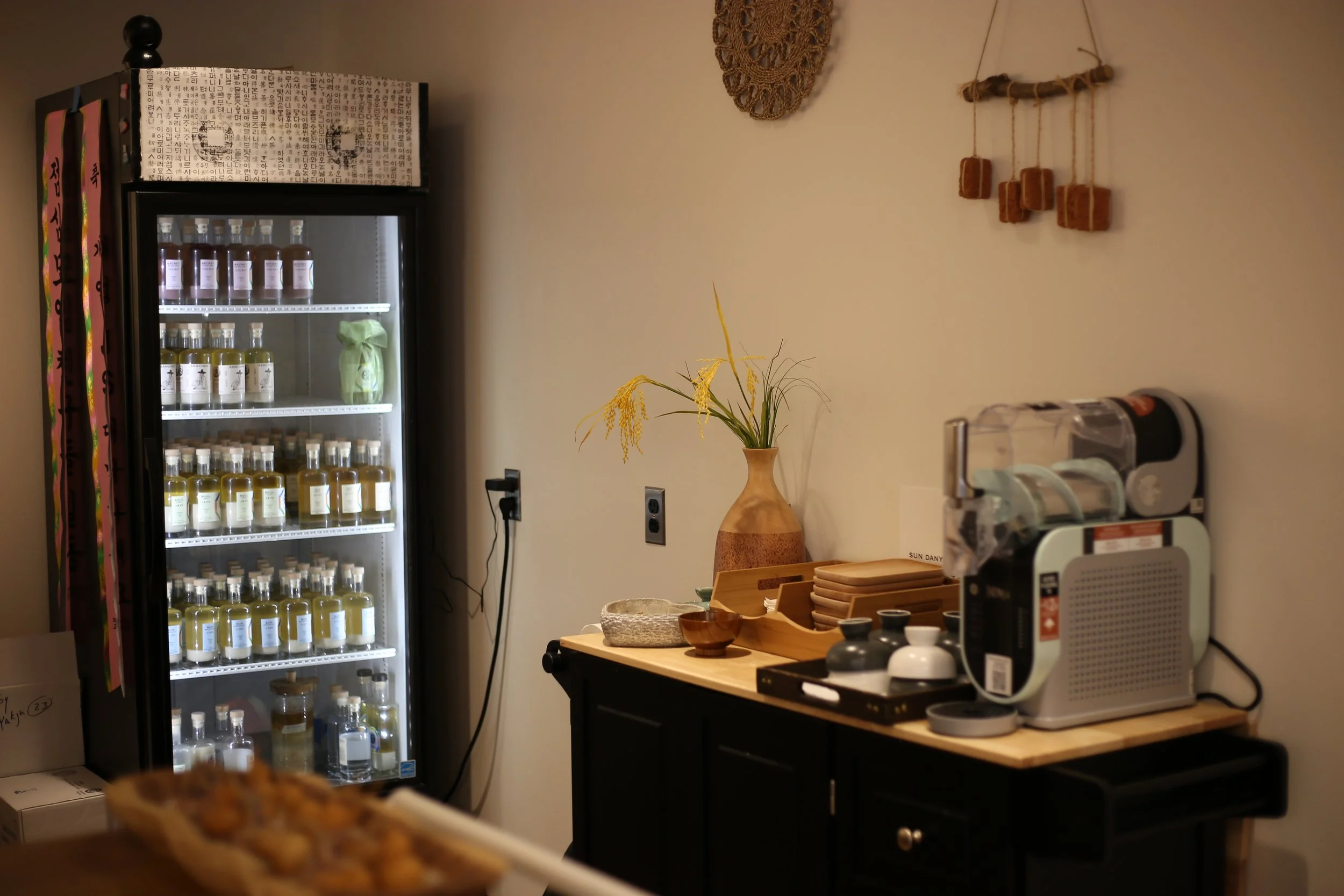 A refrigerator filled with bottles of drinks, a vase with yellow flowers on a black cabinet, and a coffee machine on a wooden counter.