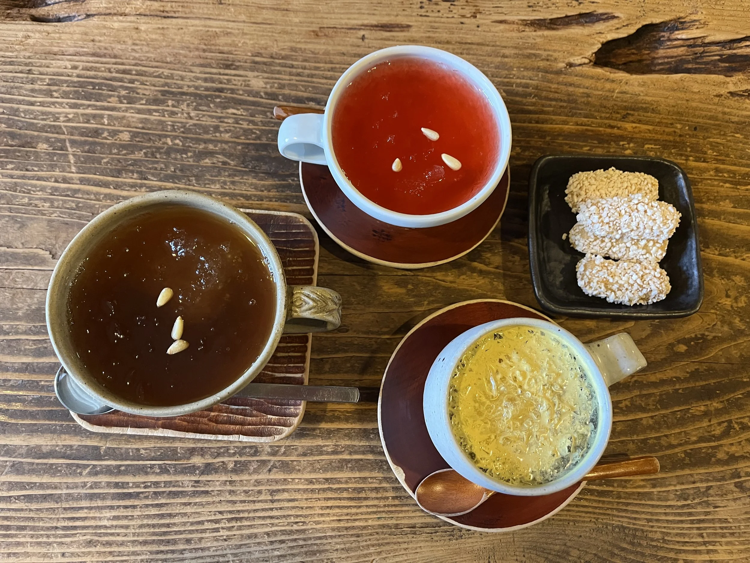 Top-down view of three mugs of different soups, a small black plate with three rice or sesame snacks, on a rustic wooden table.