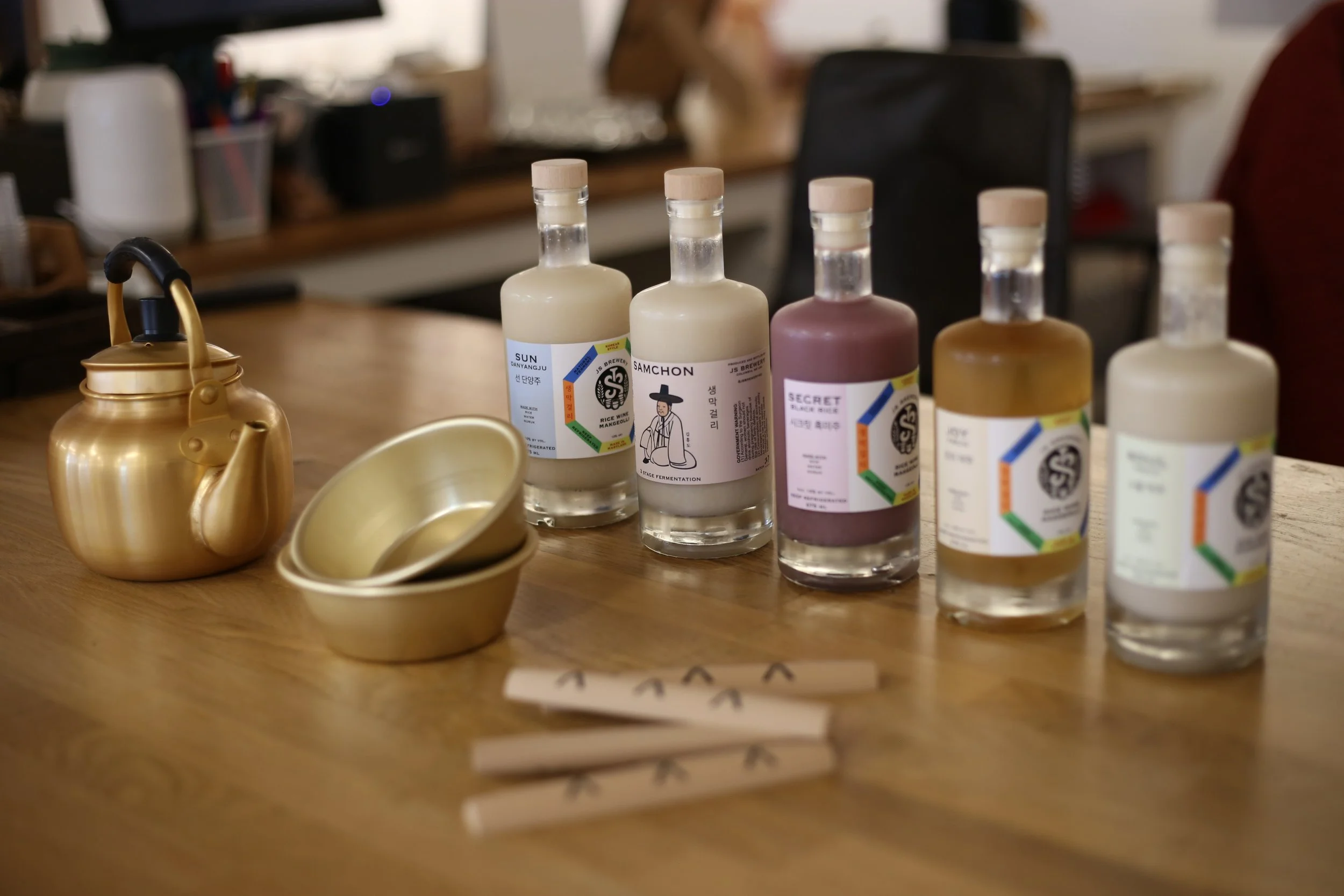 A wooden table with five bottles of sauces or dressings, stacked golden teapots and bowls, and some chopsticks with a black arrow design, in a casual dining setting.