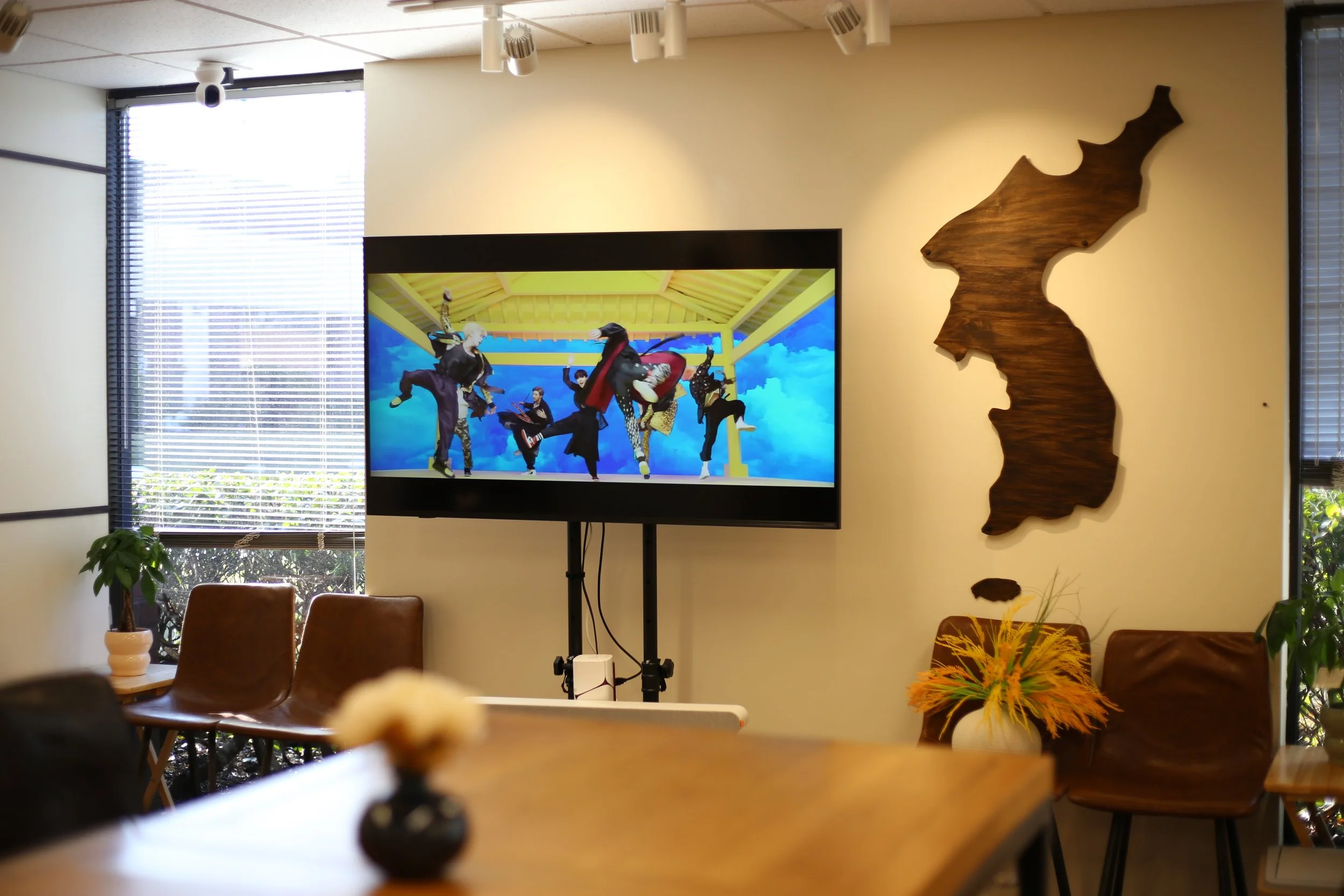 A room with a large TV screen displaying dancers in colorful costumes. The room has a wooden wall decor shaped like a map, brown chairs, a table, and potted plants.