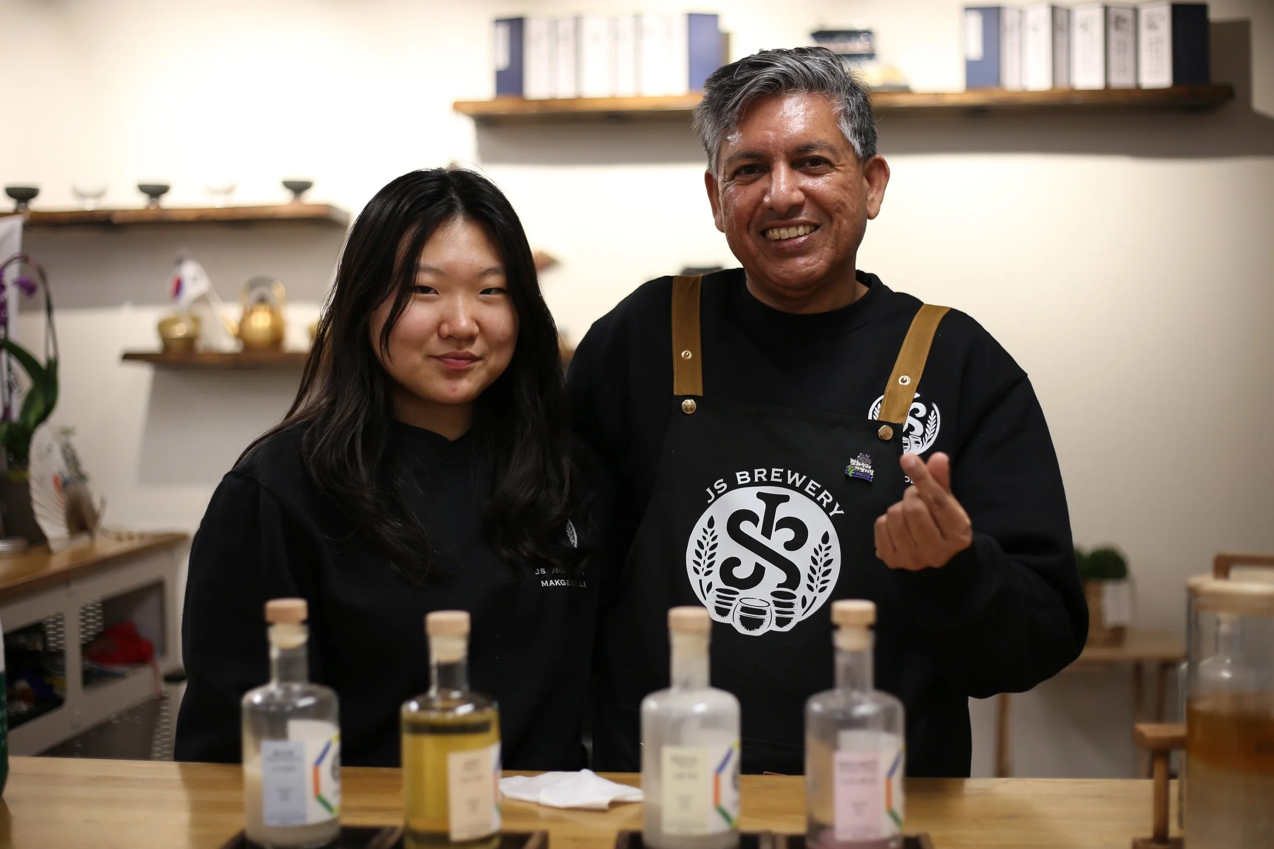 Two people smiling at the camera, one woman and one man, in a kitchen or brewery setting, with bottles of craft ingredients or products on the table in front of them, and shelves with decorative items and books in the background.