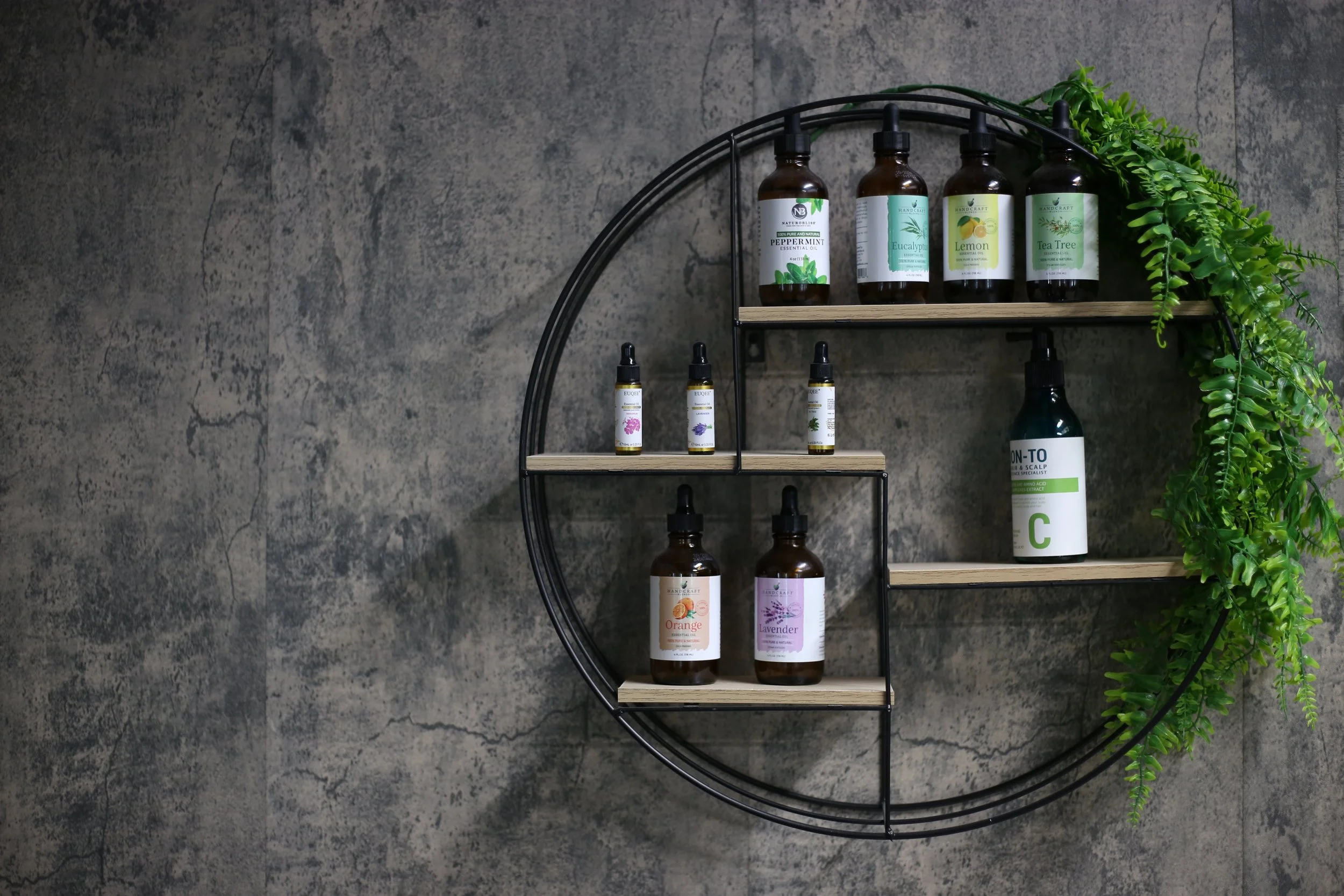 A circular wall-mounted shelf with four wooden shelves, holding essential oil bottles and a green leafy plant, against a textured gray wall.
