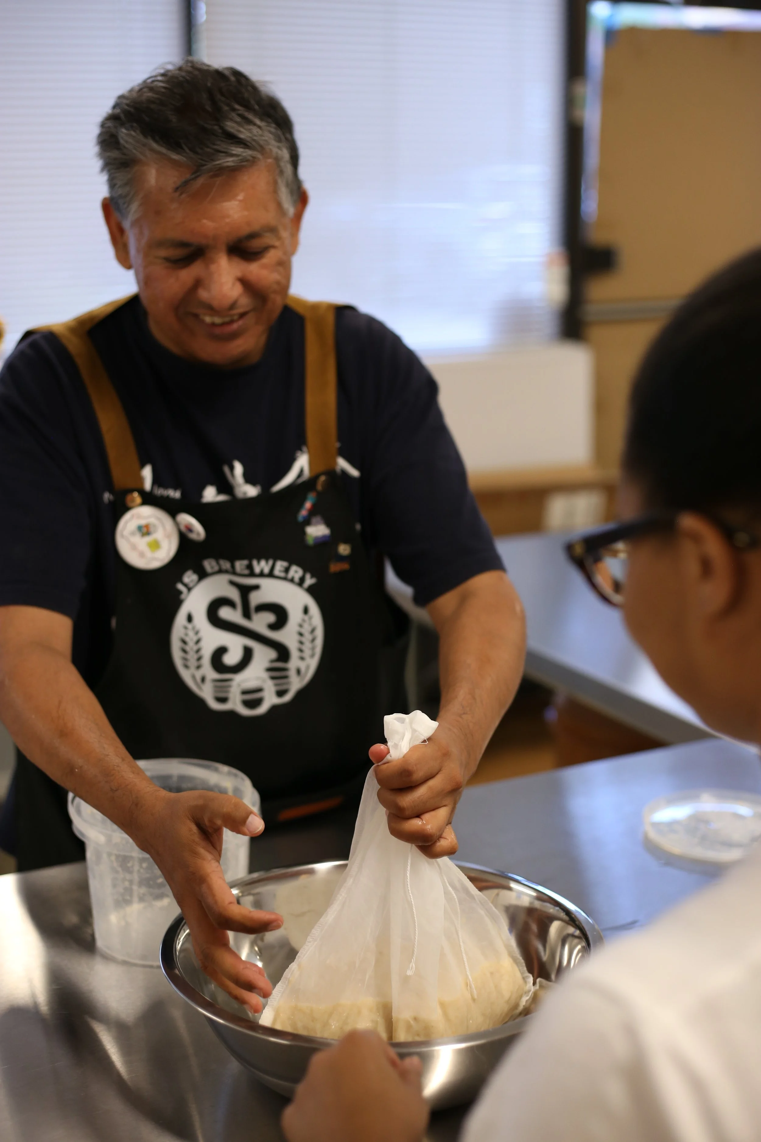 A man with gray hair wearing a black apron and buttons is stretching a large piece of dough over a stainless steel mixing bowl. There is a child with glasses assisting nearby in a kitchen or bakery setting.
