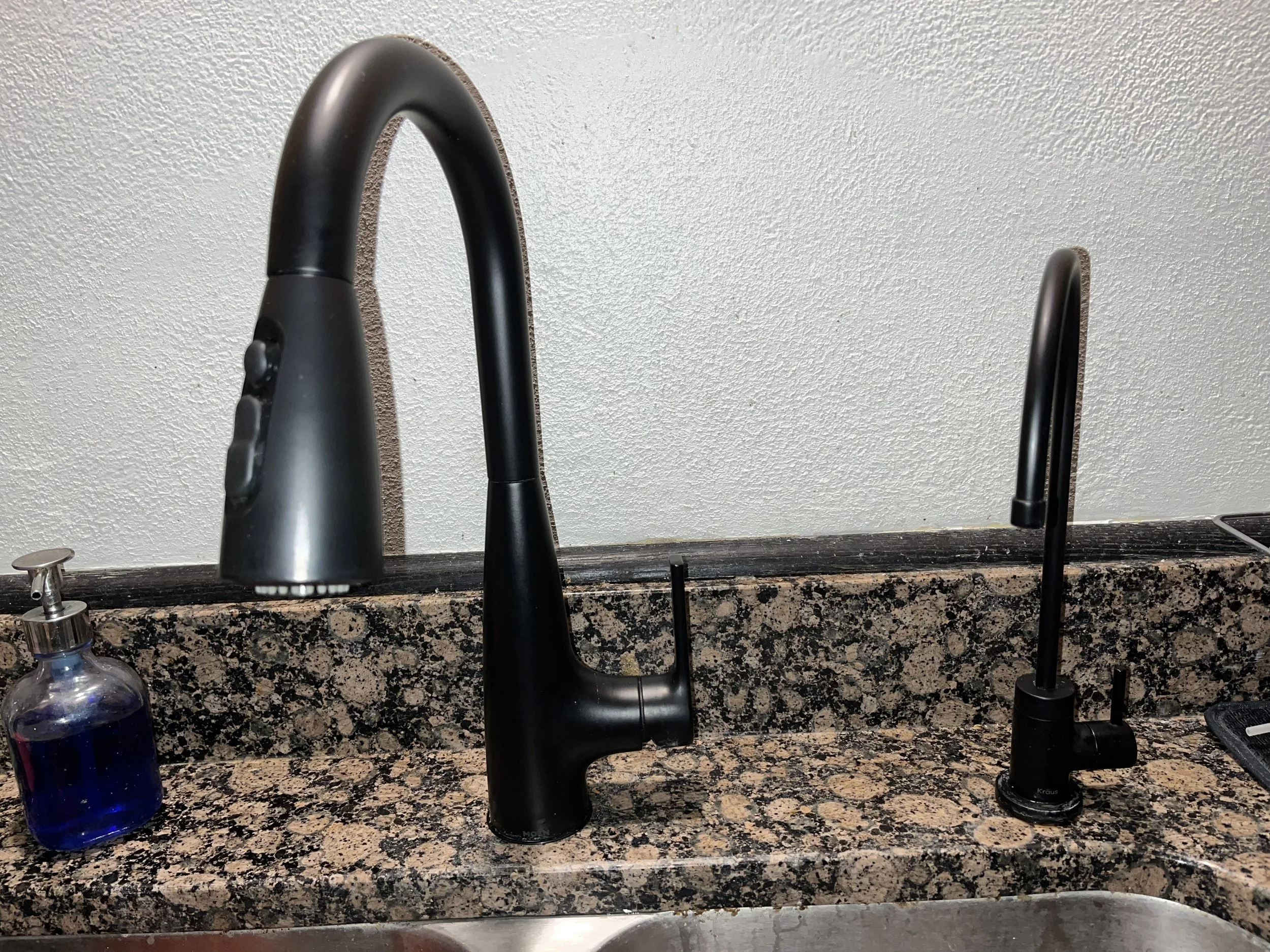 Black kitchen sink faucet and black sprayer on a granite countertop with a soap dispenser on the left and a small black object on the right.