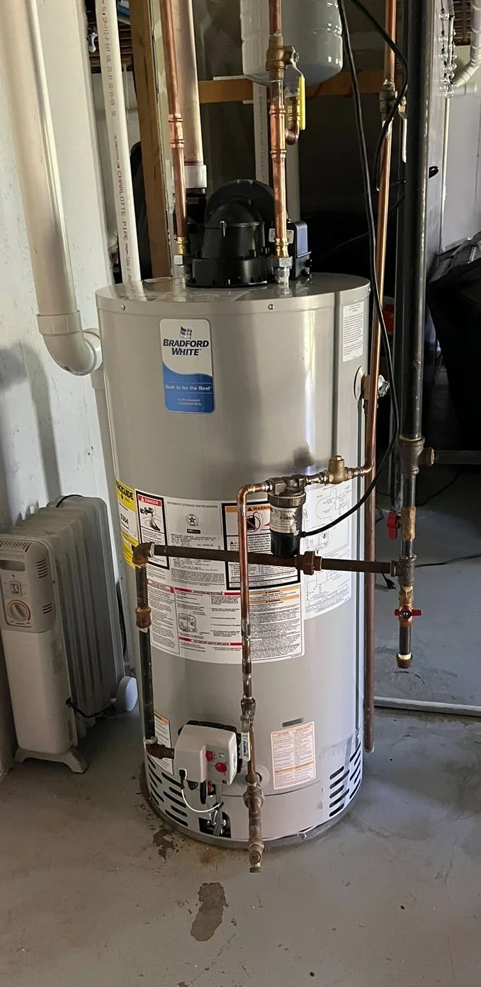 A Bradford White water heater with copper and PVC pipes connected, installed in a utility room with visible drywall and a portable space heater nearby.