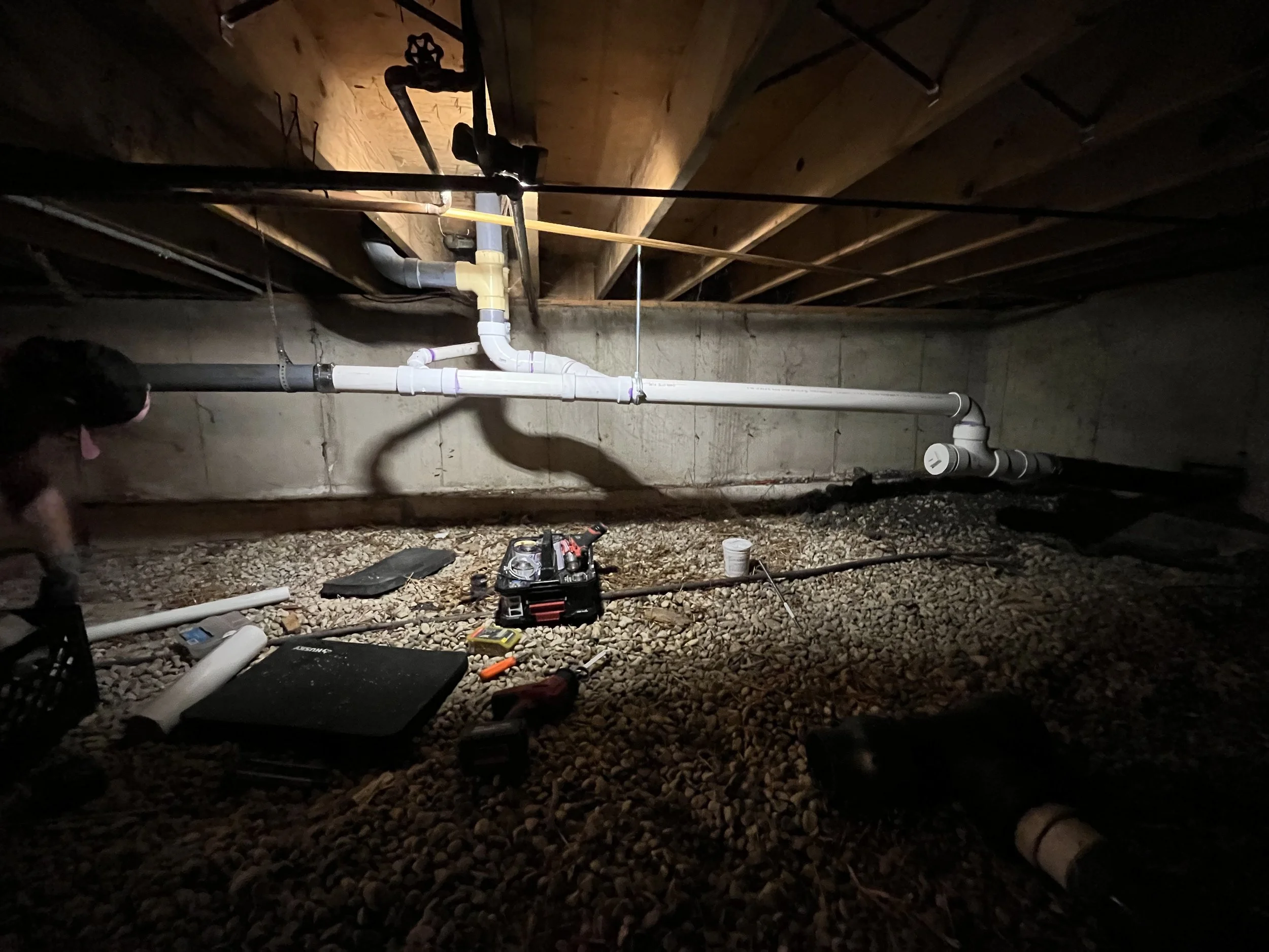 Underground space with a network of white and black pipes installed along the concrete wall, and construction tools and materials scattered on the rocky ground, including a toolbox, a cup, a drill, and a mat.