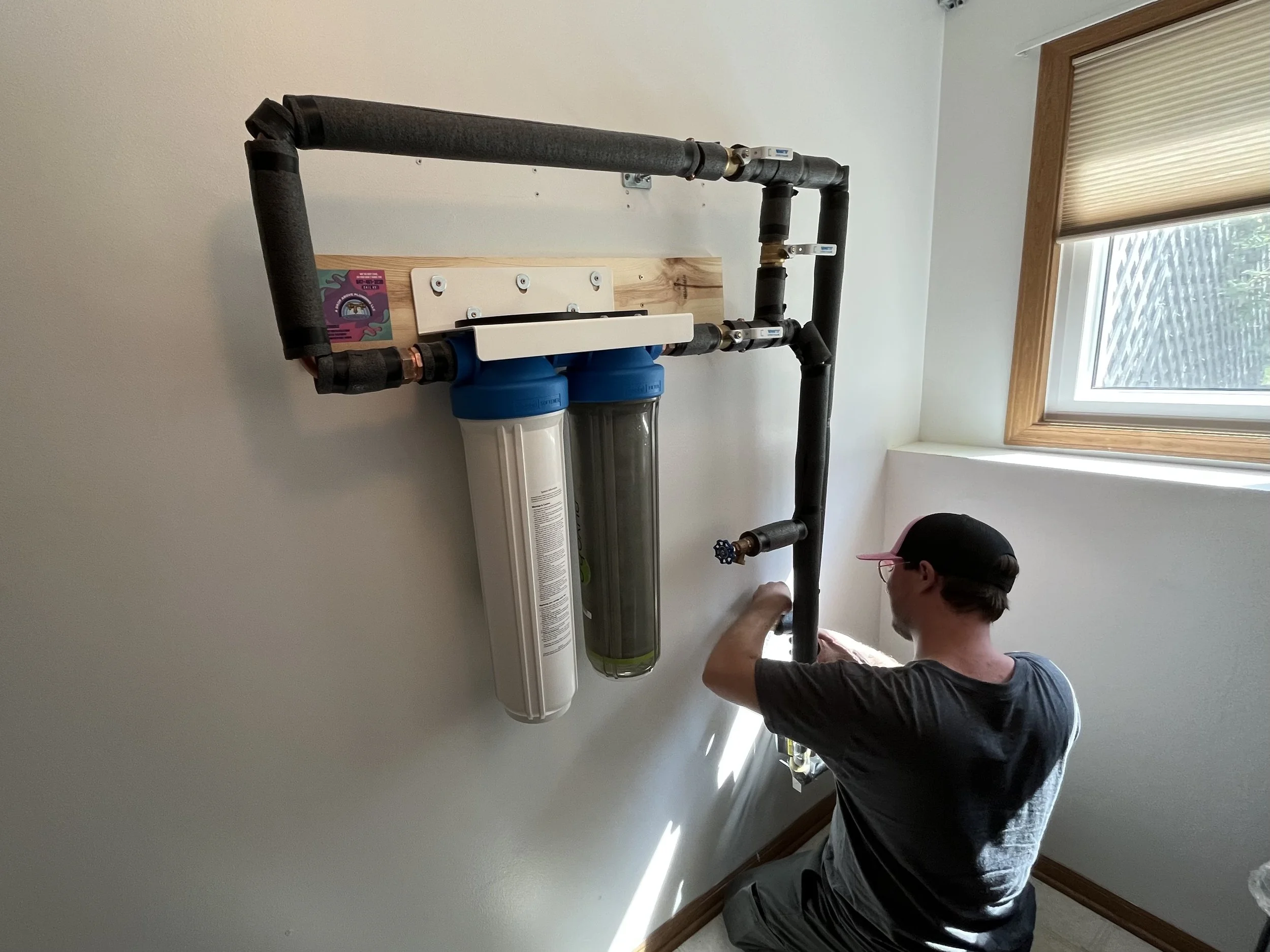 A man is installing or working on a water filtration or softening system on a wall in a room with a window. The system includes black pipes, a white filter, and a clear filter tank, with the man adjusting components.