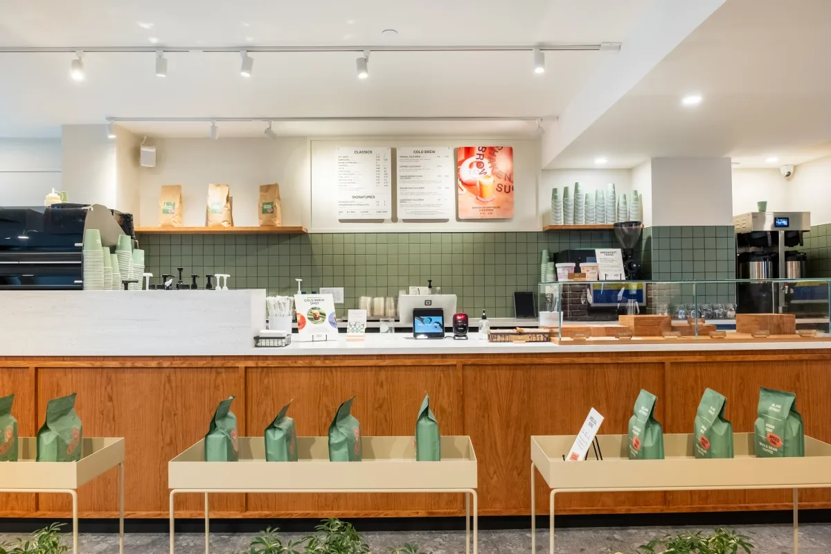 Inside a coffee shop with a wooden counter, green tiled backsplash, and shelves with coffee bags and cups. Menu boards are on the wall, and green coffee bags are displayed on tables in front.