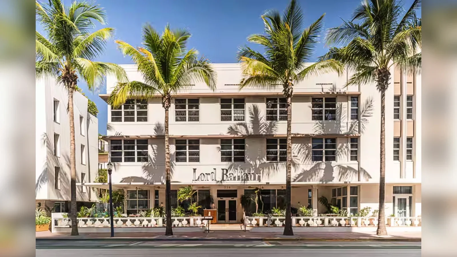 A white multi-story hotel called Lord Basinger with large windows, surrounded by tall palm trees and a sidewalk in front.
