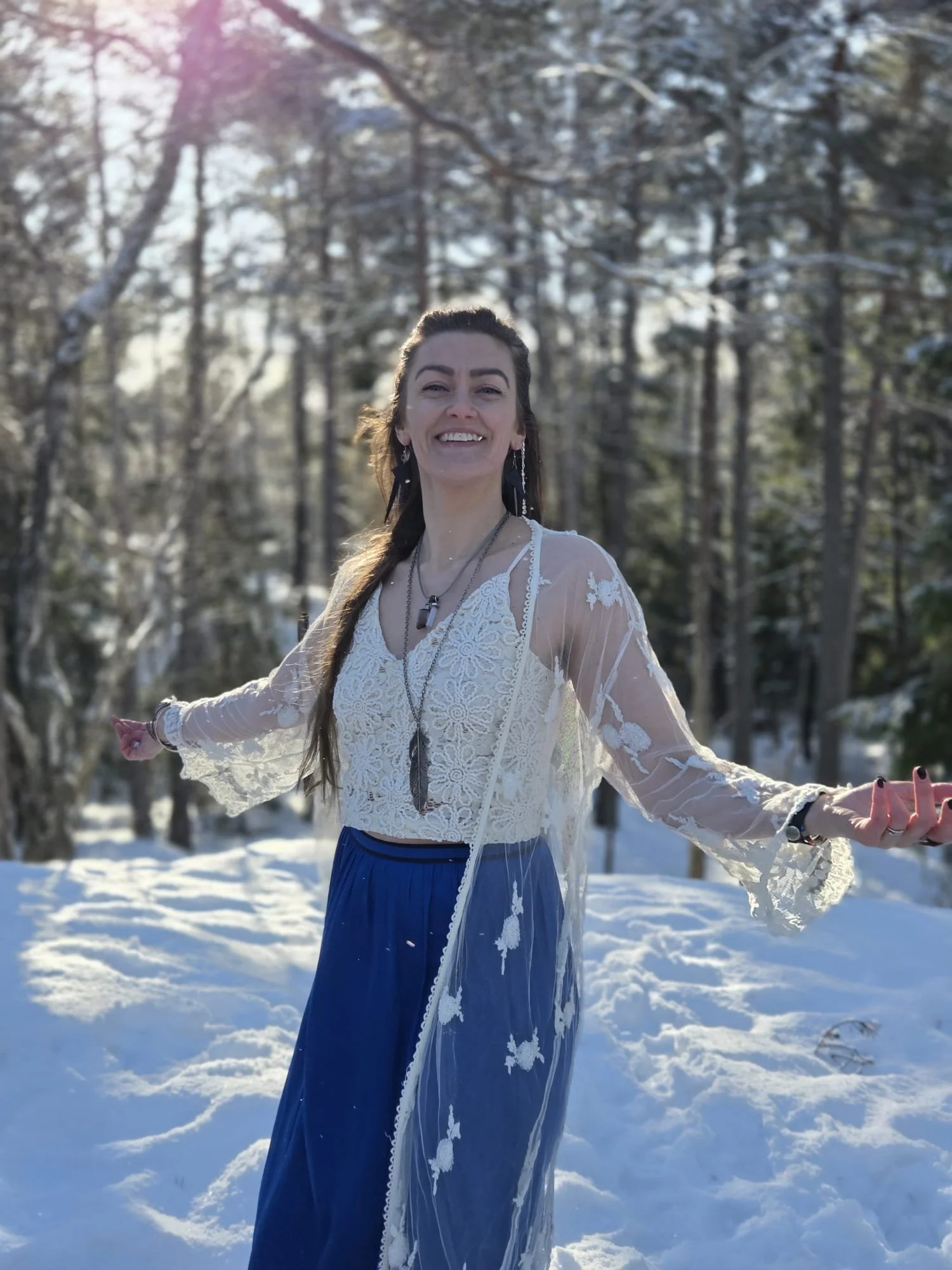A woman standing in a snow-covered wooded area, smiling with open arms, wearing a white lace top and a long blue skirt, with sunlight filtering through the trees.