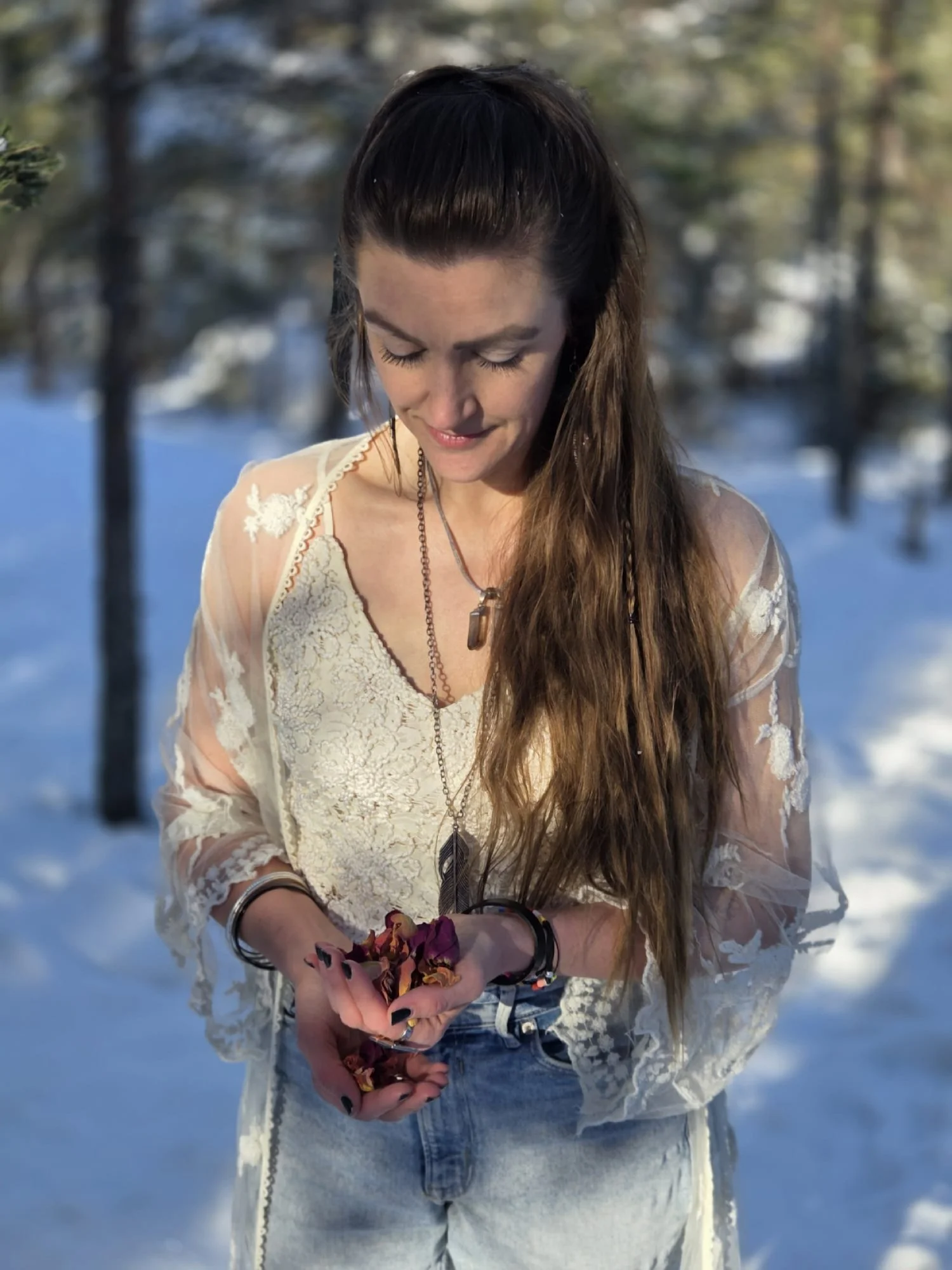 A woman with long brown hair, wearing a white lace blouse and denim jeans, stands outdoors in a snowy forest during the daytime, holding dried rose petals in her hands and looking down with a gentle smile.