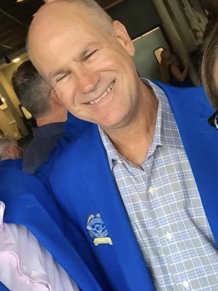 Percy Legendre, wearing a blue jacket and a checked shirt, standing in an indoor setting.
