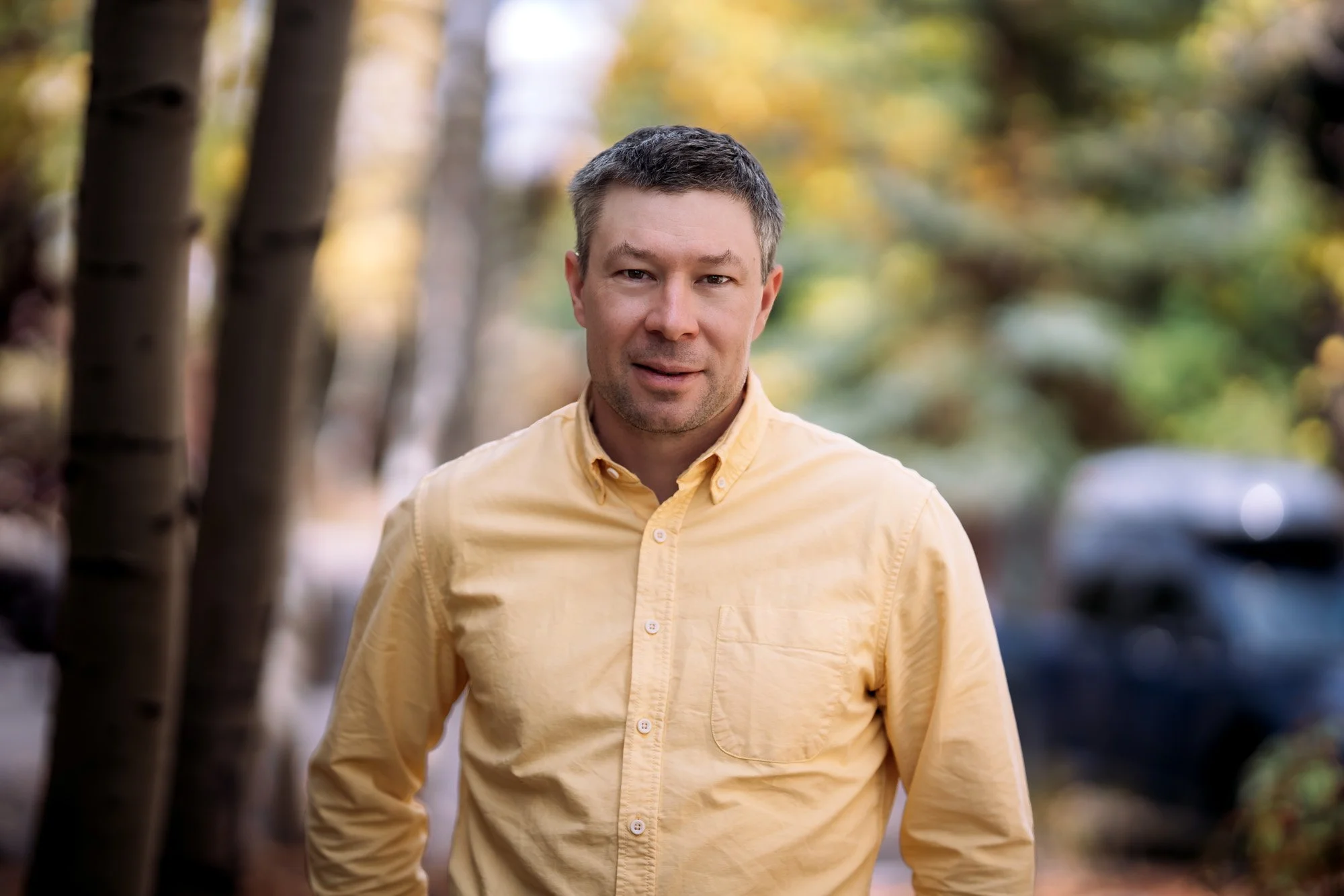 A man with short brown hair and a light stubble beard, wearing a yellow button-up shirt, standing outdoors with blurred autumnal trees and parked cars in the background.