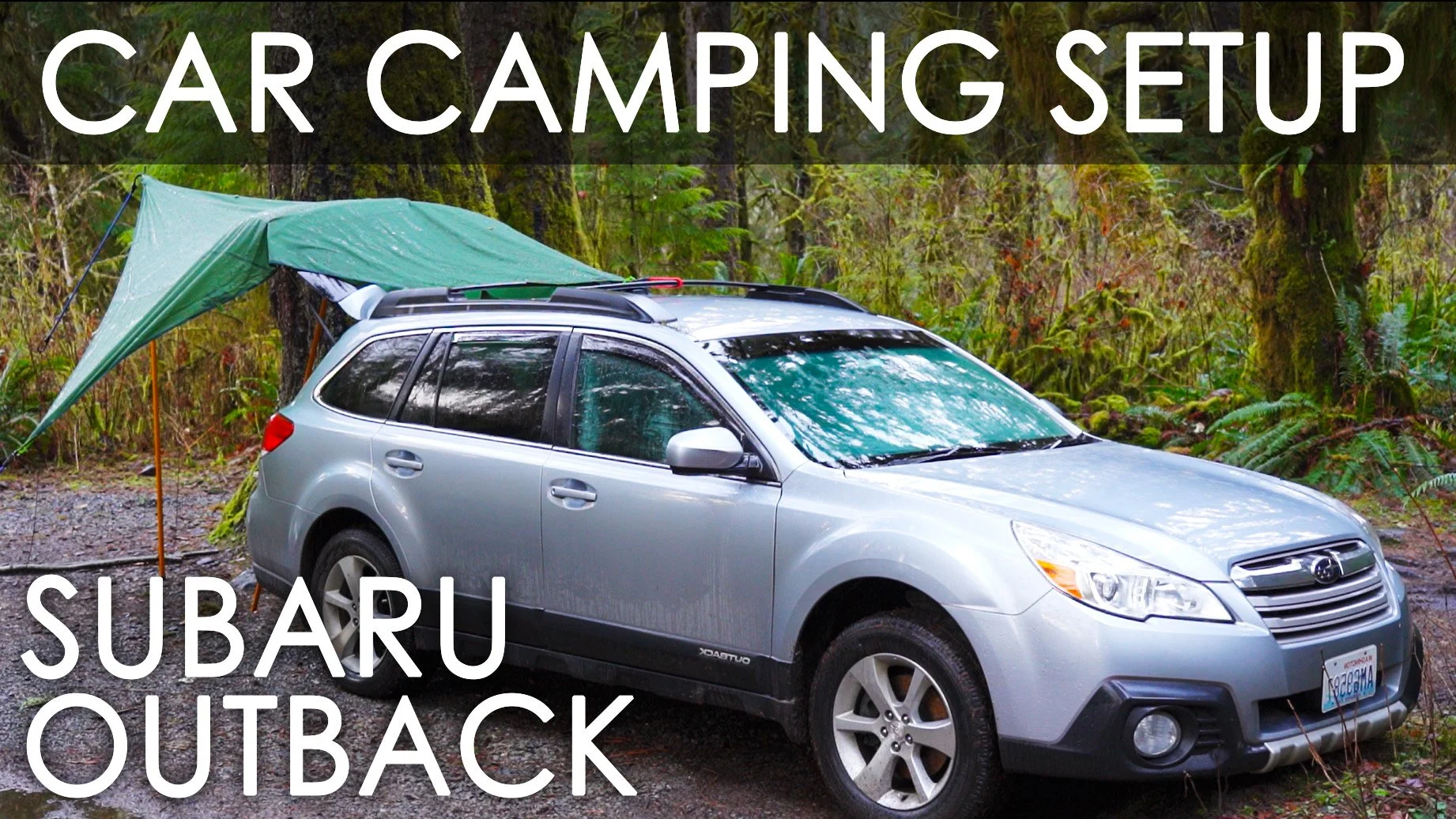 Silver Subaru Outback parked in a wooded area with a green tarp tent attached to the roof rack, surrounded by green trees and foliage.