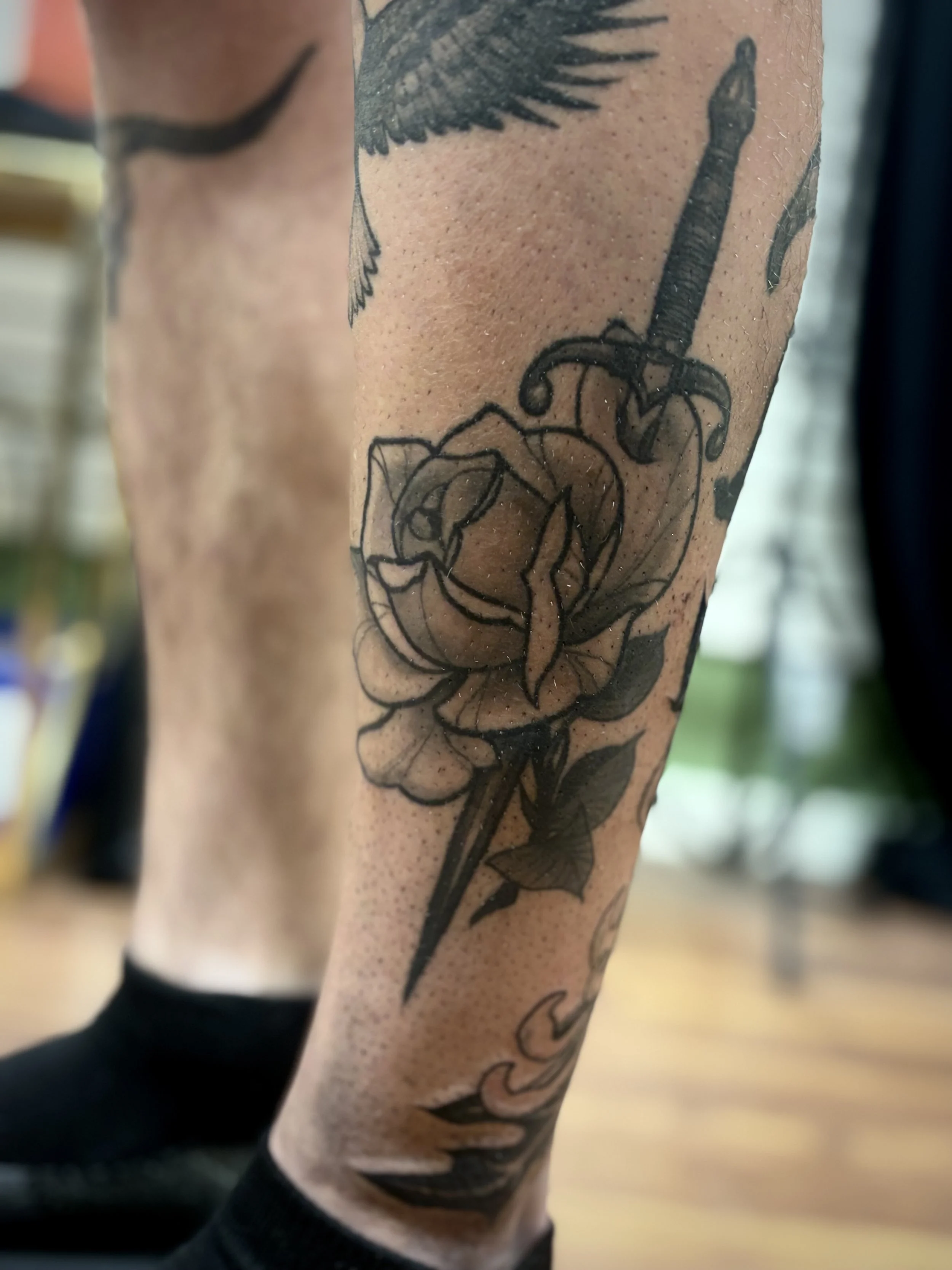 A black and grey tattoo of a rose with a dagger through it on a person's leg.