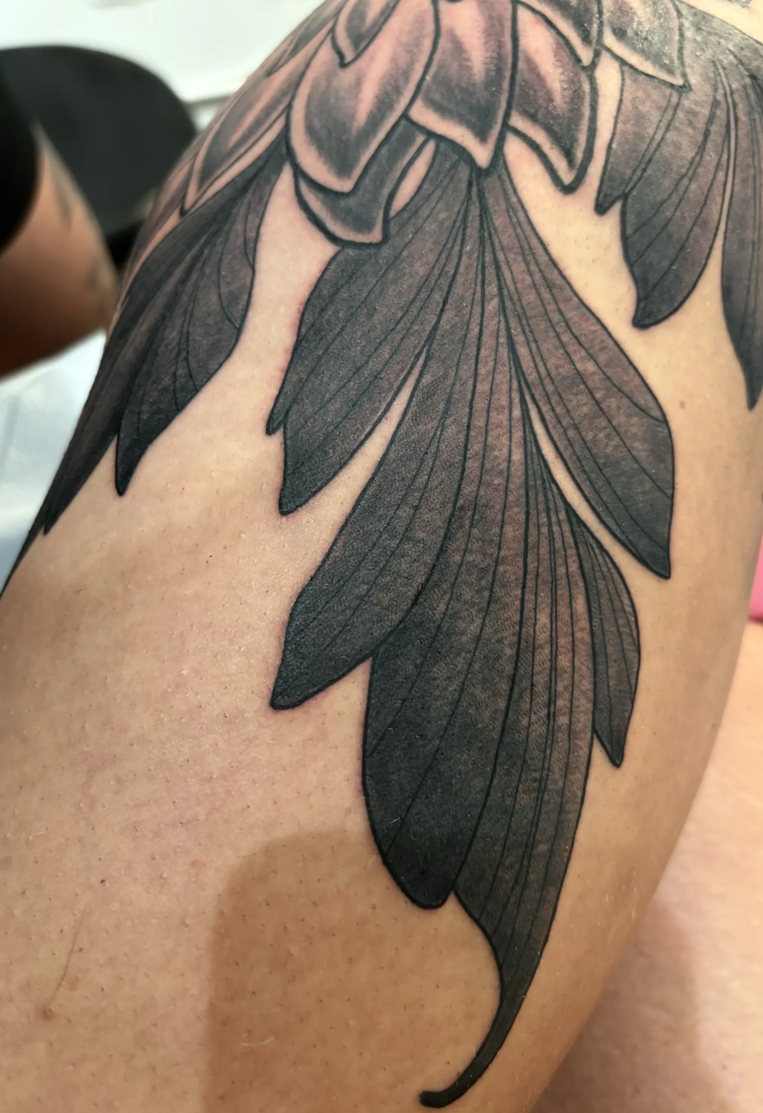 Close-up of a black ink floral tattoo on a person's shoulder, featuring large petals with detailed line work.
