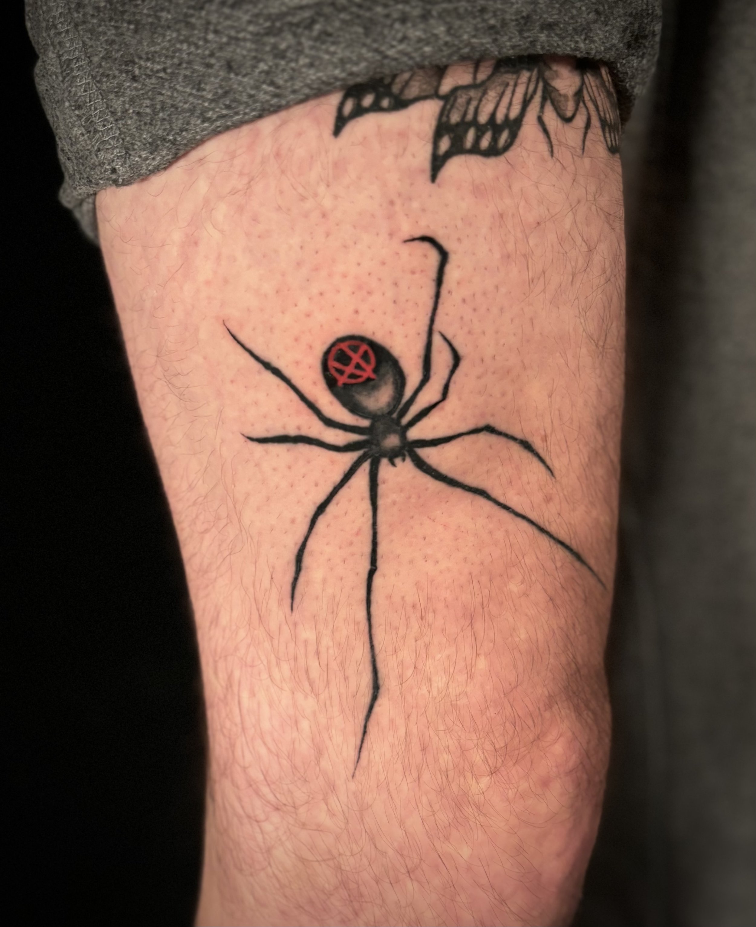 Tattoo of a black widow spider with a red upside-down cross on its abdomen on a person's arm.
