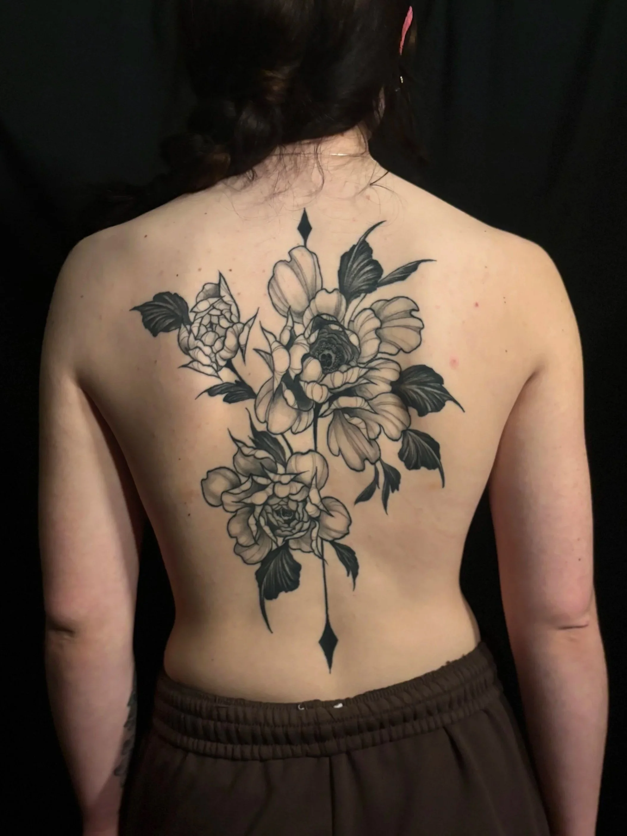 Black and grey back tattoo of a large, detailed black and gray floral design with peonies, leaves, and decorative elements on a person's back.