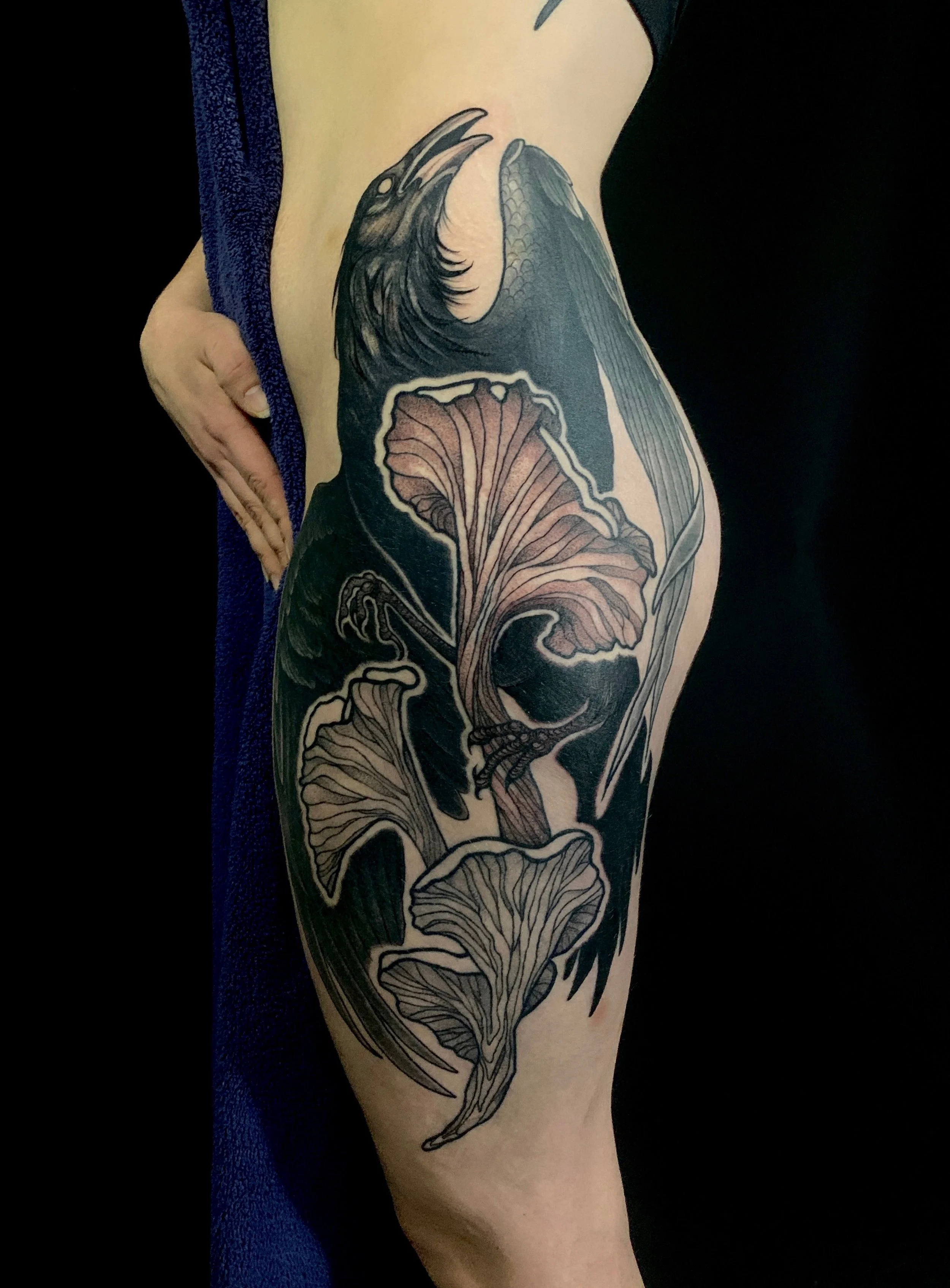 Tattoo of a black and gray raven and large mushrooms with intricate details on a person's thigh.