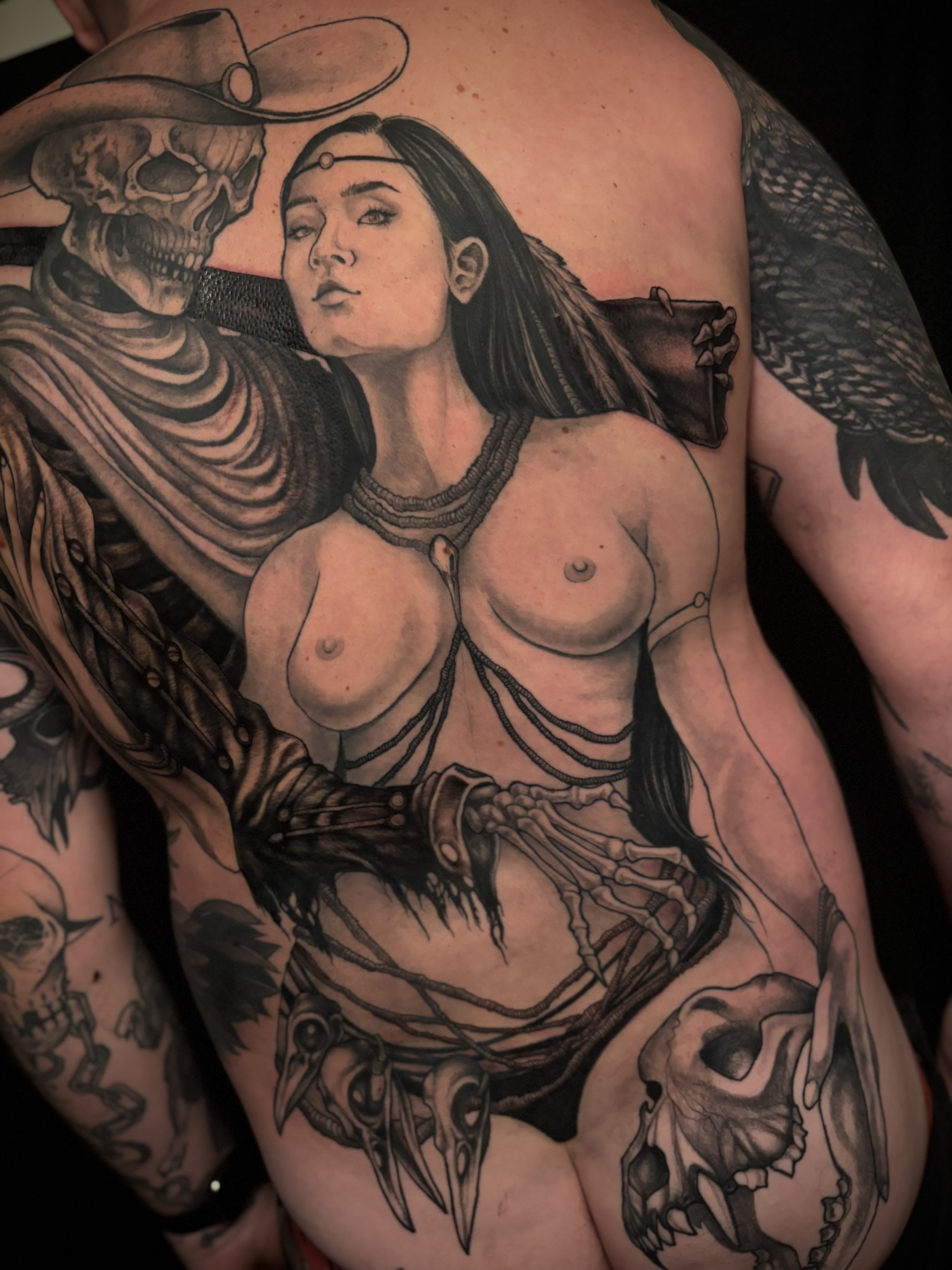 A detailed black-and-gray tattoo of a First Nations woman with long hair, wearing jewelry, holding a skeleton wearing a cowboy hat, and with skulls and bones at the bottom.