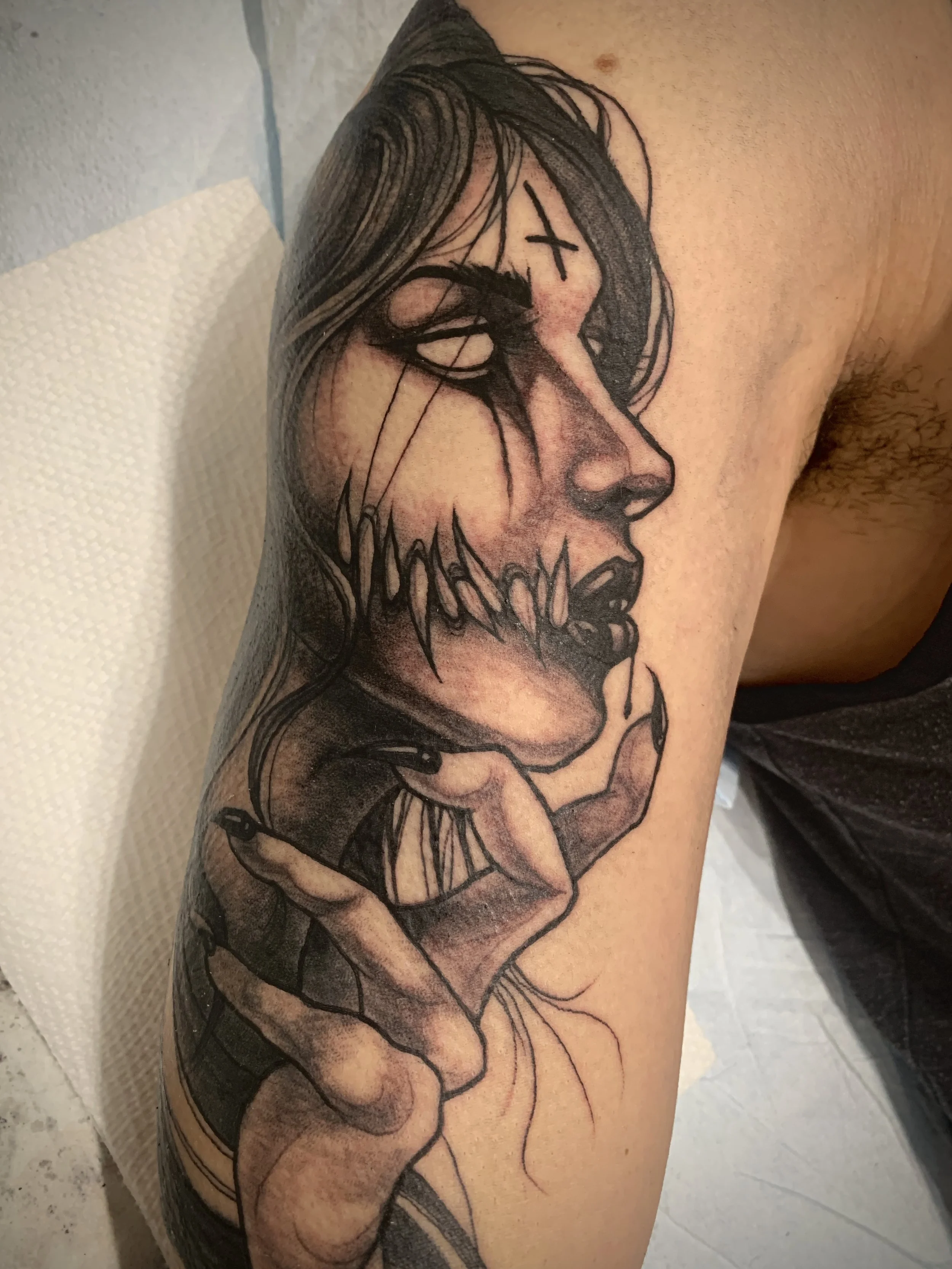 Black and gray tattoo of a demon woman's profile with vacant eyes, and a large demonic mouth with teeth spreading up her cheek and long flowing hair, holding her hand near her chin, detailed with shading and line work.
