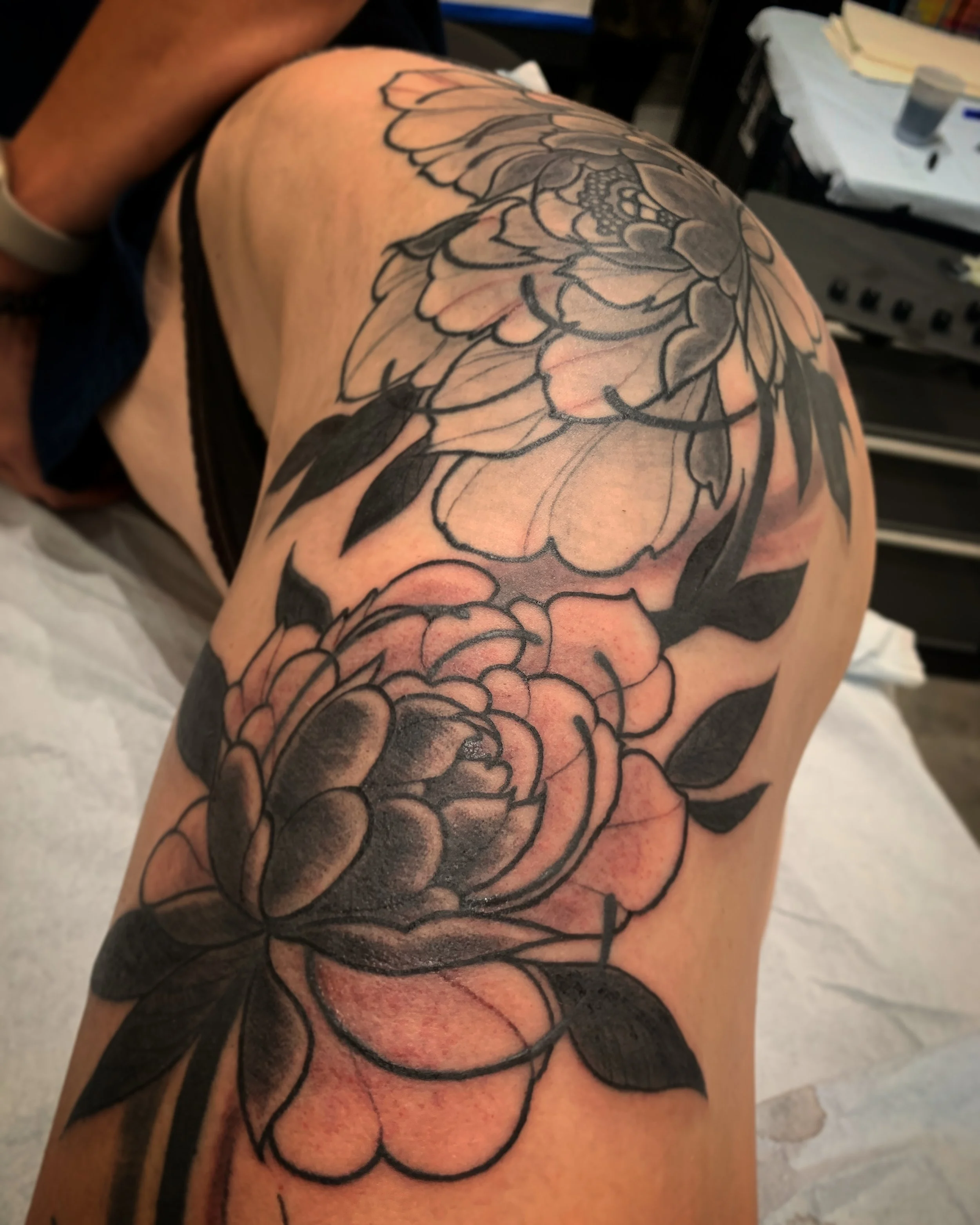 A person's thigh with a black and gray floral tattoo featuring large peony flowers, leaves, and detailed line work.