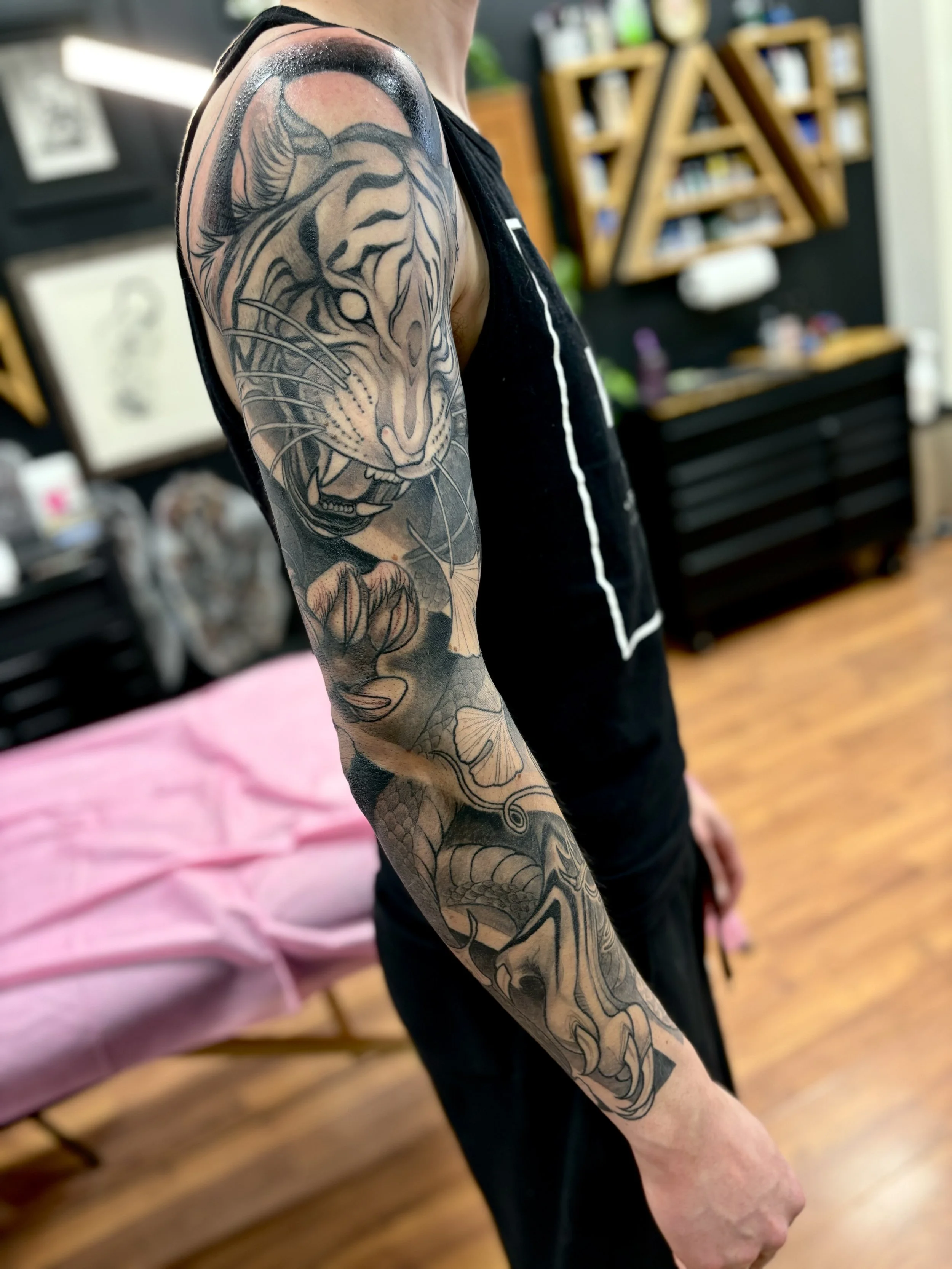 Close-up of a person's right arm with a detailed black and grey tattoo of a tiger, snake, and gingko leaves sleeve, in a tattoo studio.
