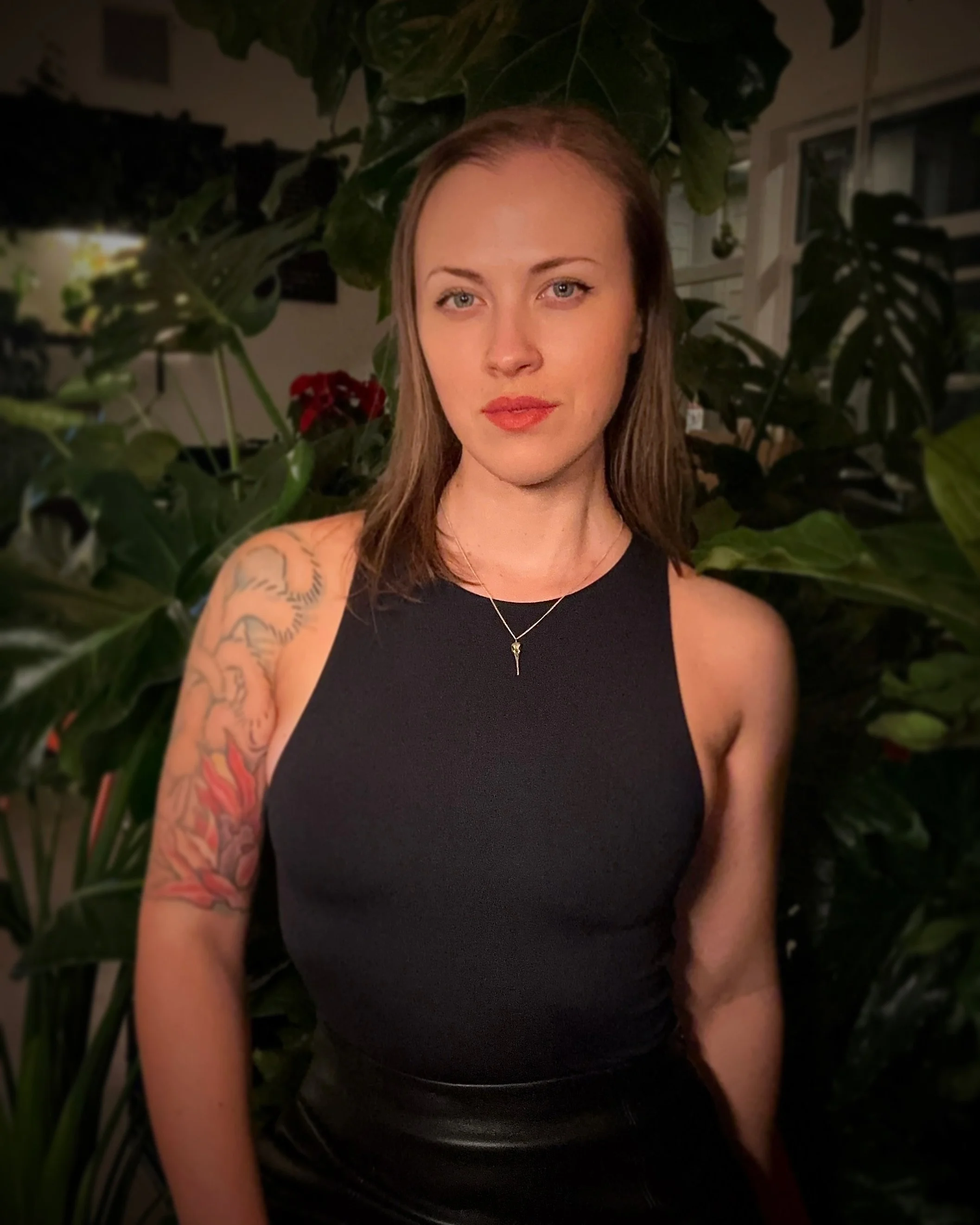 Portrait of tattoo artist Sarah Chalmers, dressed in black. There are many dark, glossy plants in the background framing her.