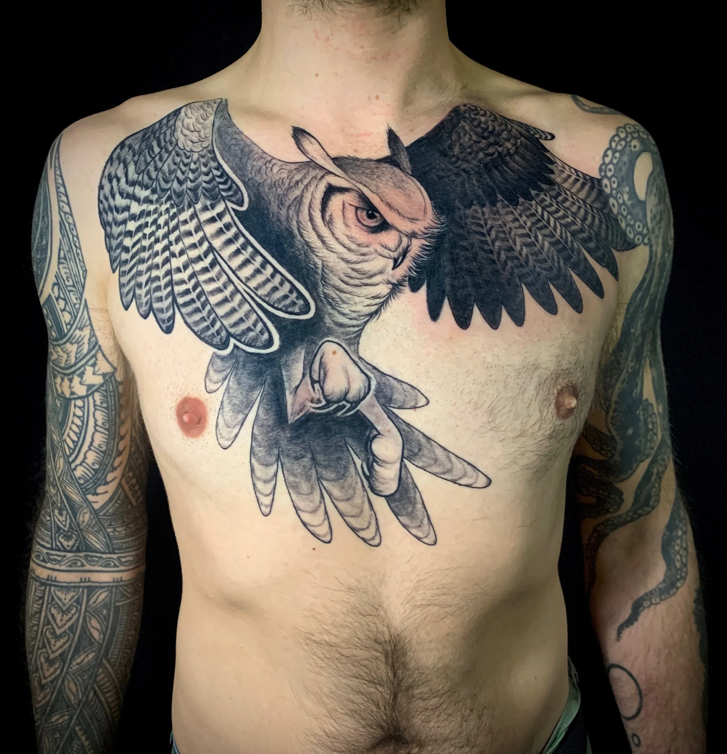 A person with extensive chest and arm tattoos, featuring a large great-horned owl with detailed feathers on his chest and extending down past the sternum.