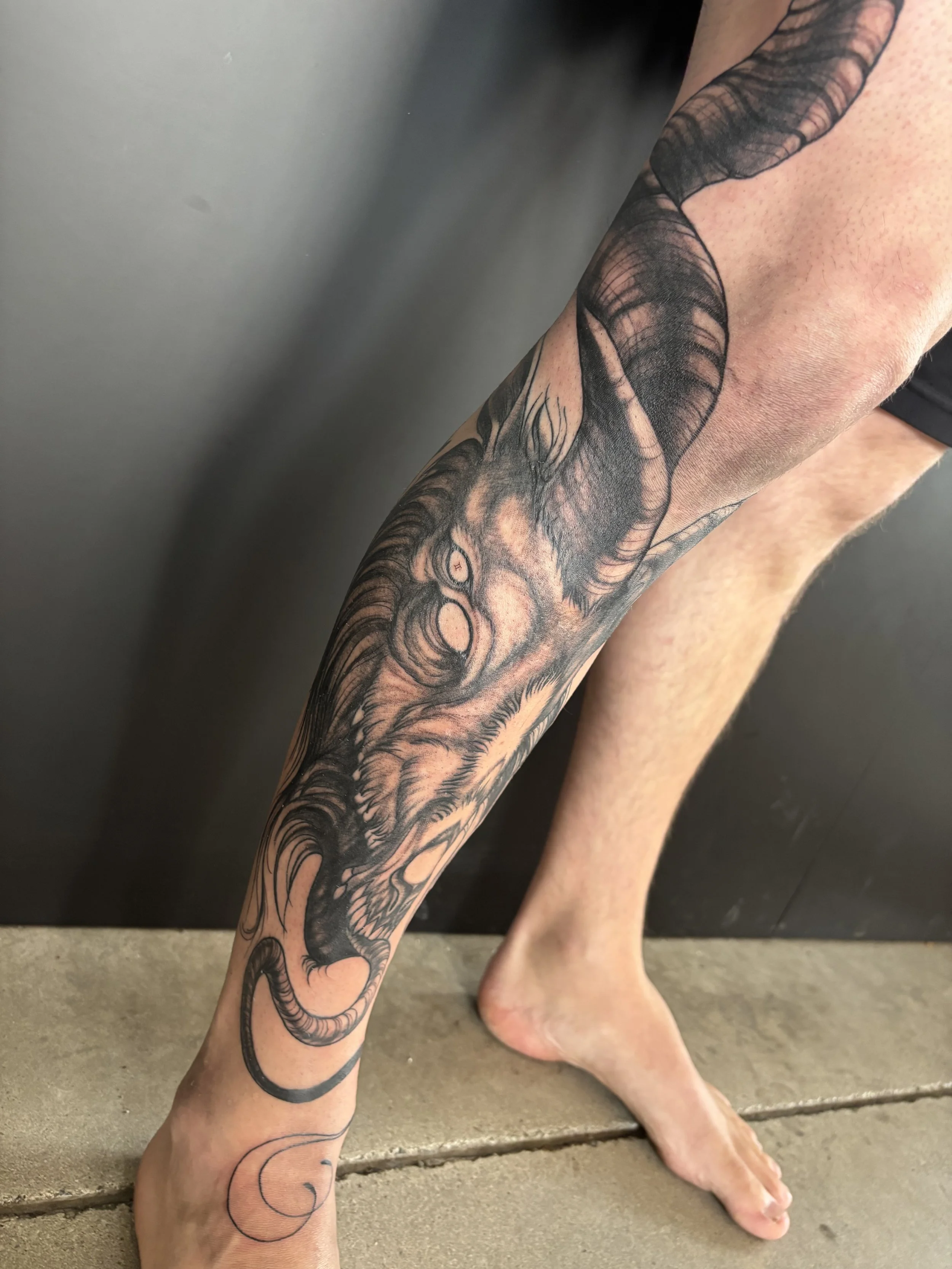 A person's lower leg features a large, detailed tattoo of a demonic goat face with prominent horns and fierce eyes, on a person's leg with its horns going up the persons thigh.
