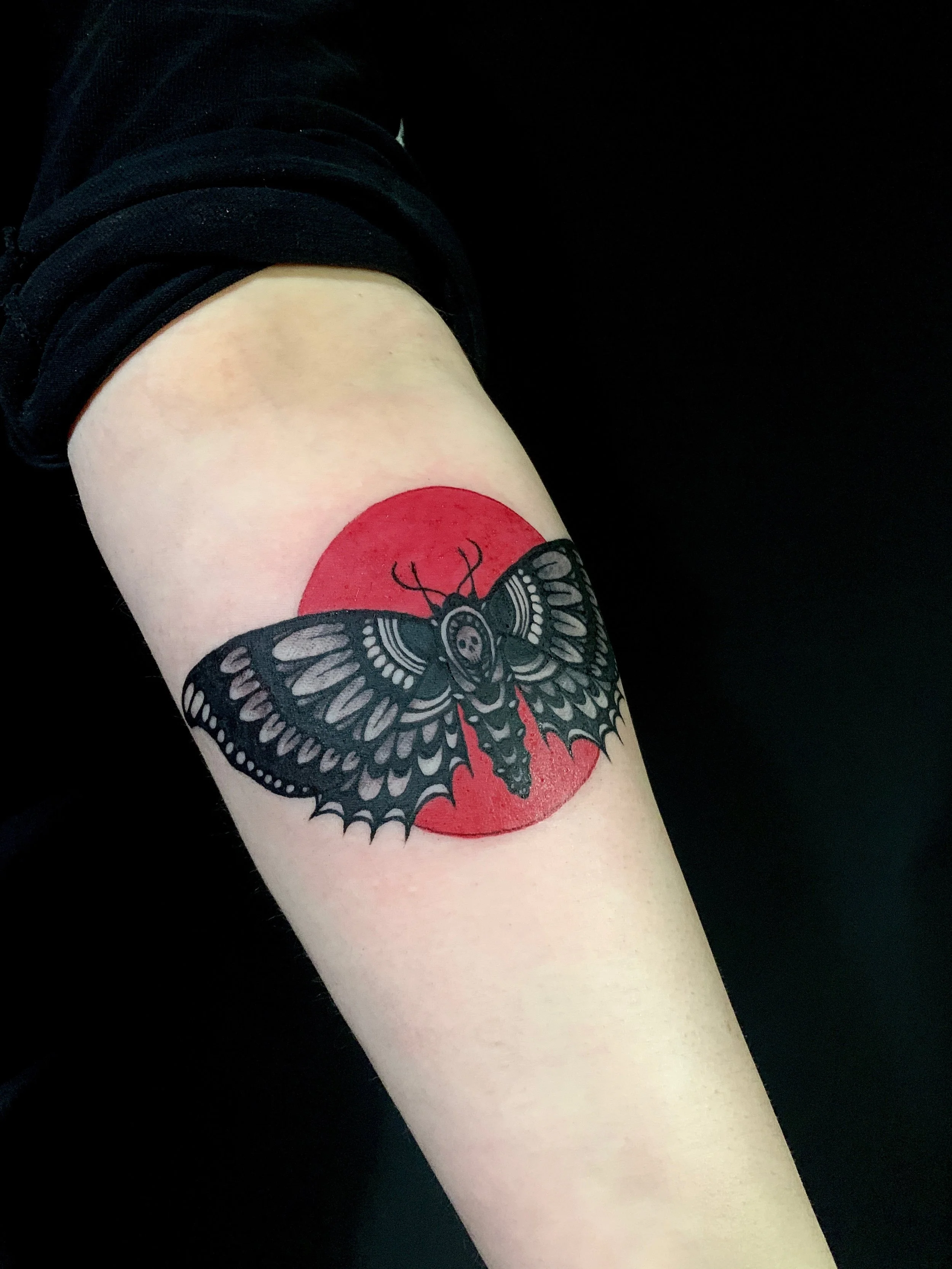 Tattoo of a moth with black and gray details on a person's arm, with a red circle background.
