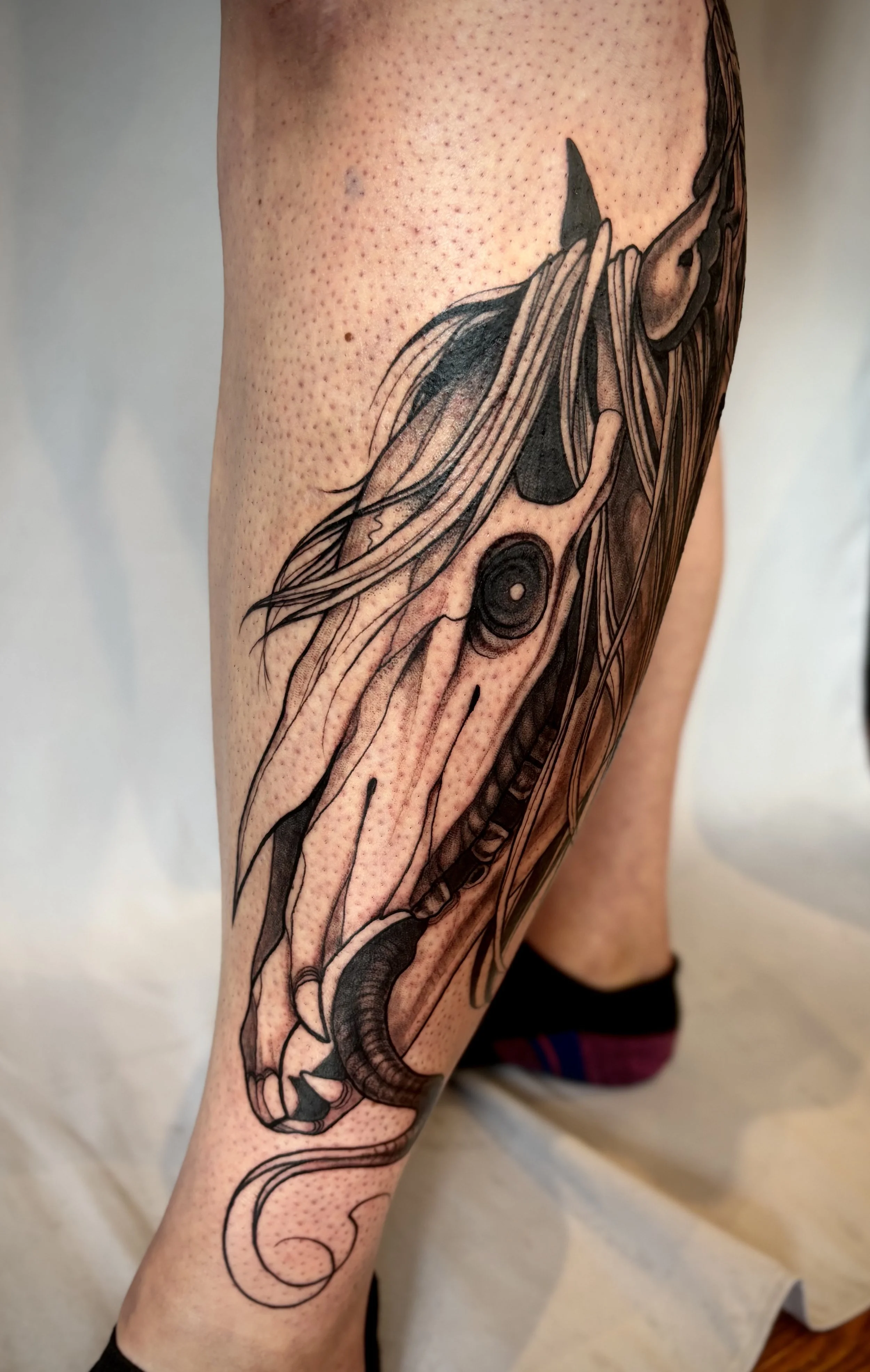 Black and gray tattoo of a horse skull on a person's calf, showing a detailed mane, long demonic tongue and detailed facial features.