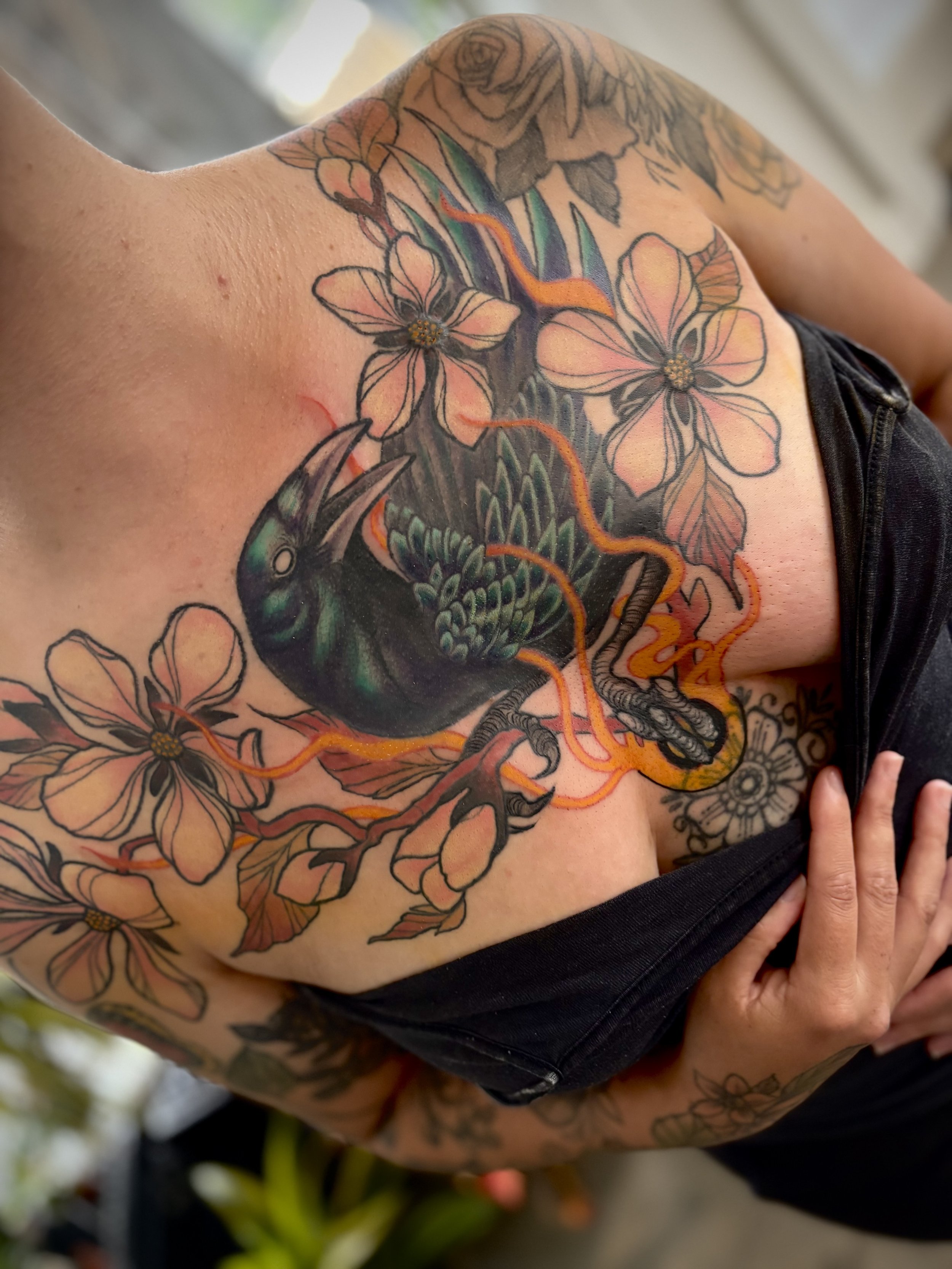 Colorful tattoo of cherry blossoms, a black crow, fiery orange and yellow flames, and a black floral design on a person's chest.